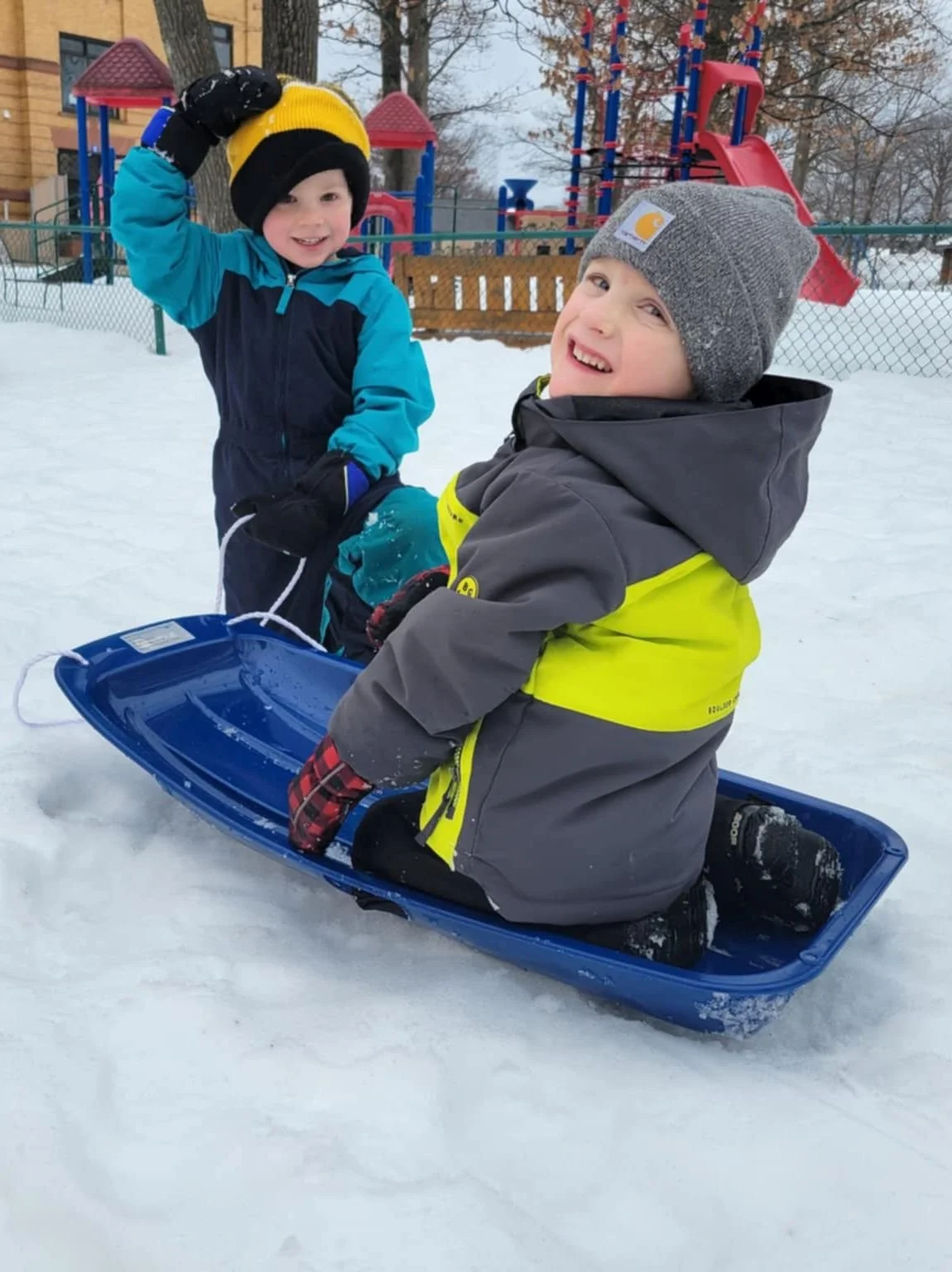 Close up of 2 preschool students outside on a winter day.  One student is on his knees on a blue sled in the snow.  The  other  student is kneeling in the snow in at the front of the sled, holding the chords that pull the sled.