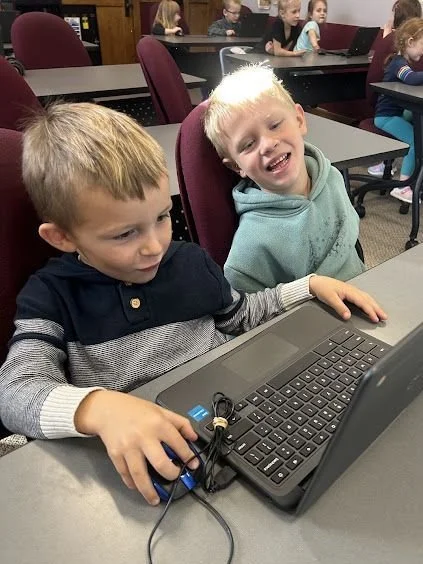 Two kindergarten students, smiling, sitting in front of an open Chromebook.