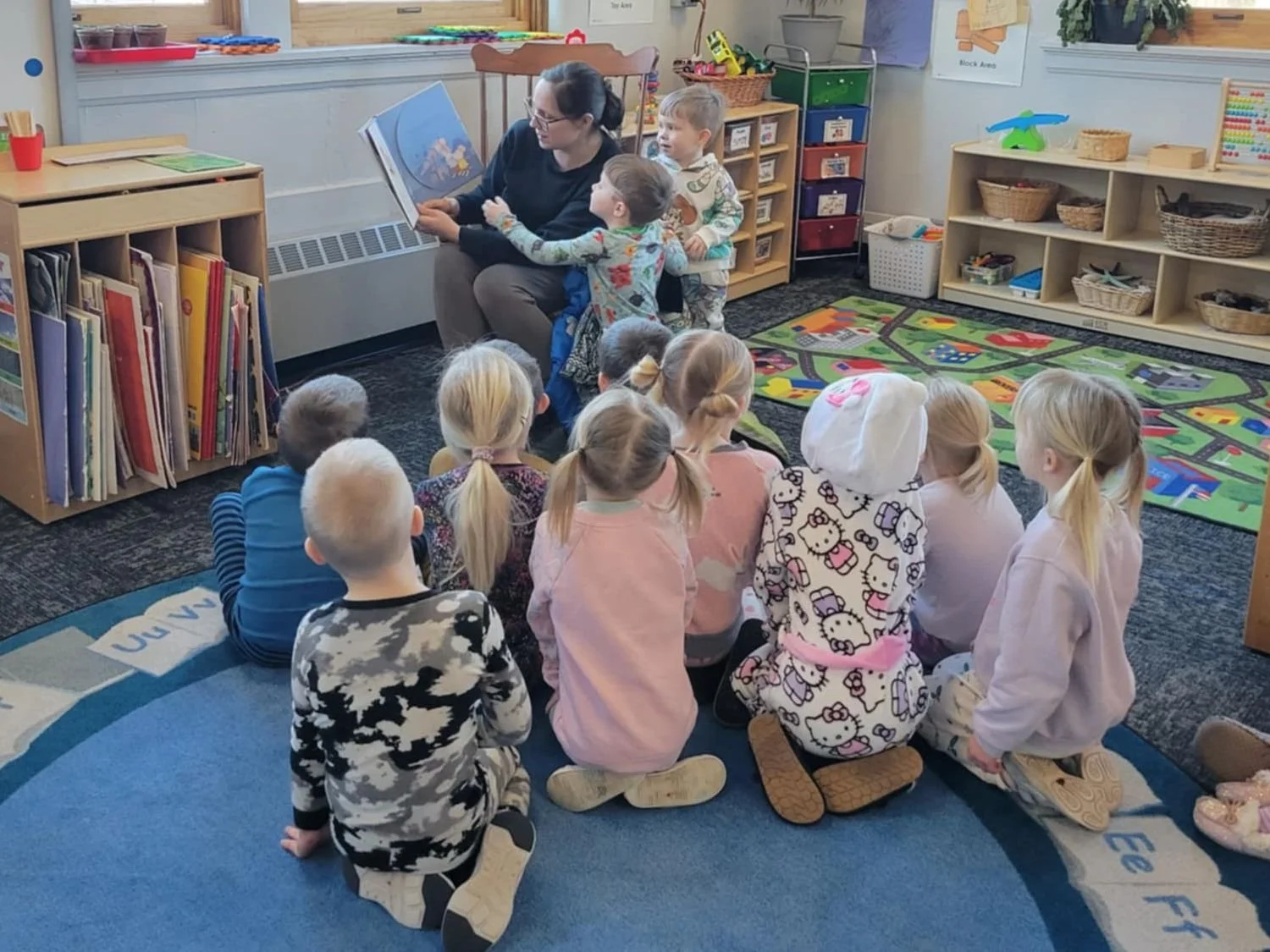 In the background is an assistant sitting in a rocking chair facing the class and reading a book.  In the foreground is the back of the heads of a dozen preschool children attentively listening to the story.