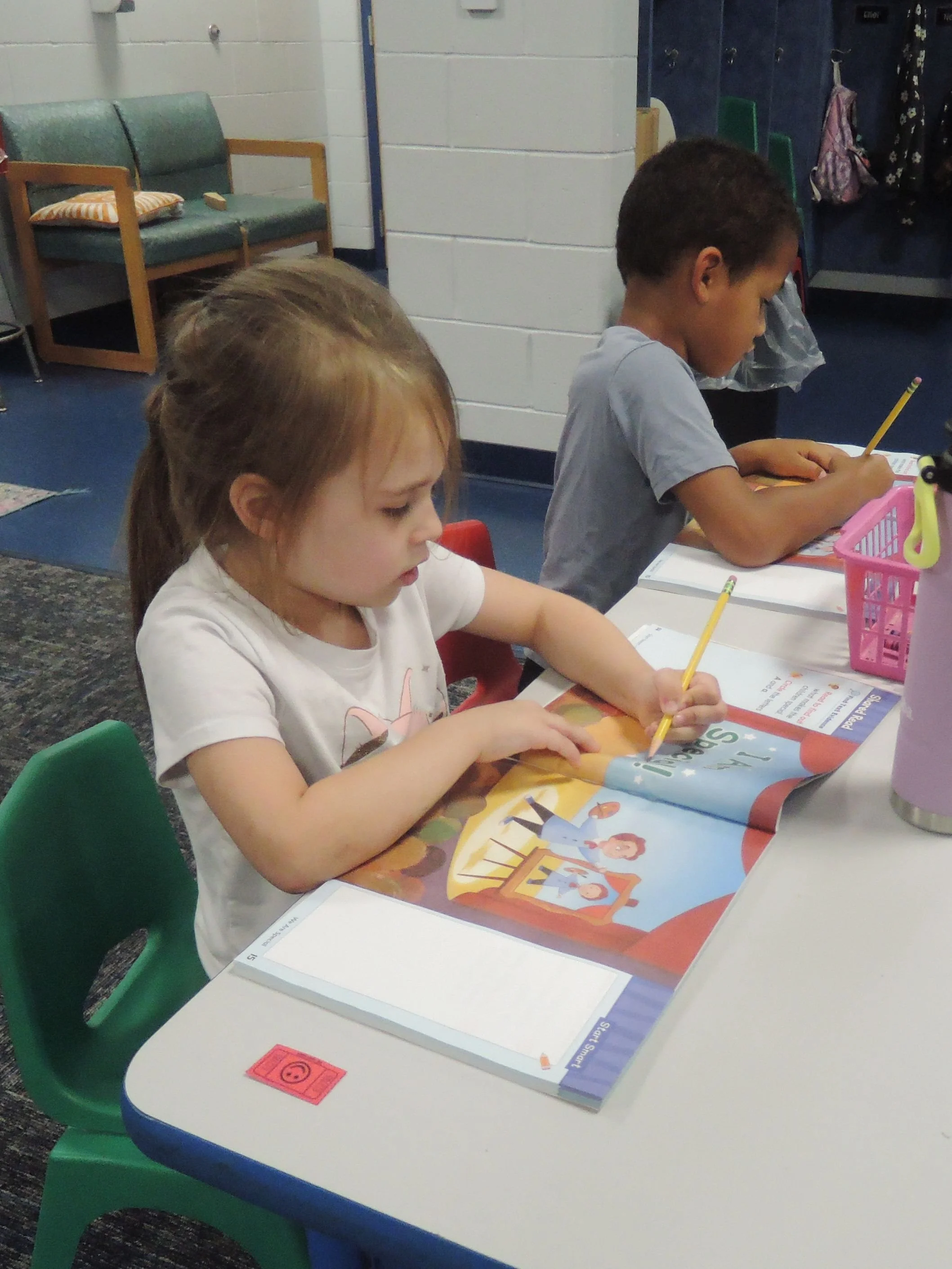A side view of two kindergarent students sitting at their shared desk.  They booth have colorful workbooks open and pencils in hand as they work intently on an assignment.