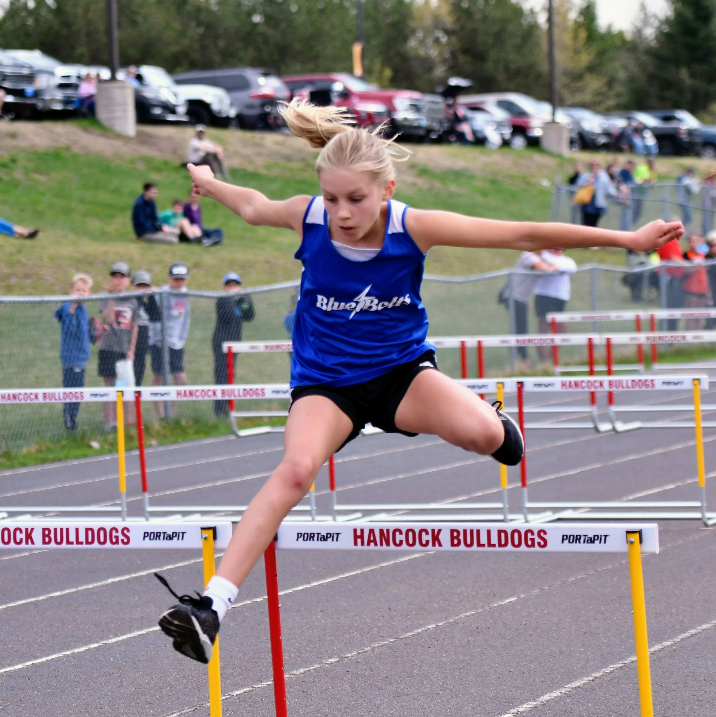 A junior high girl in blue top and black shorts in mid jump over a hurdle.