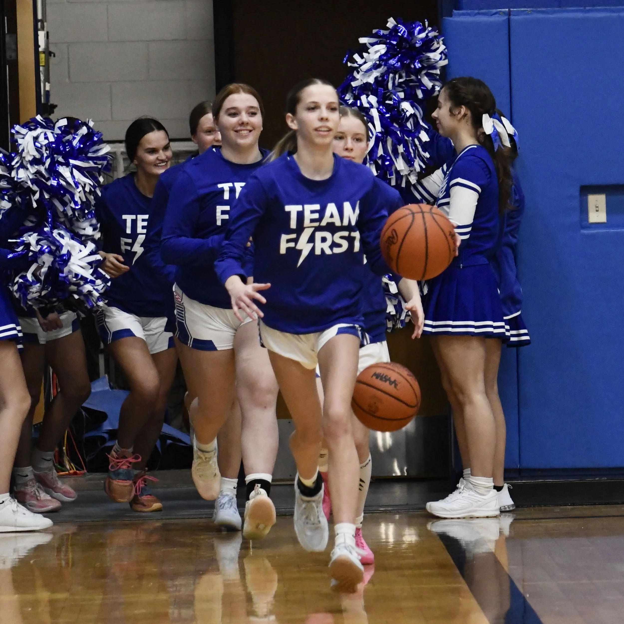 Varsity players racing on to the court single file, with cheerleaders on both sides  using pom poms to cheer them on.