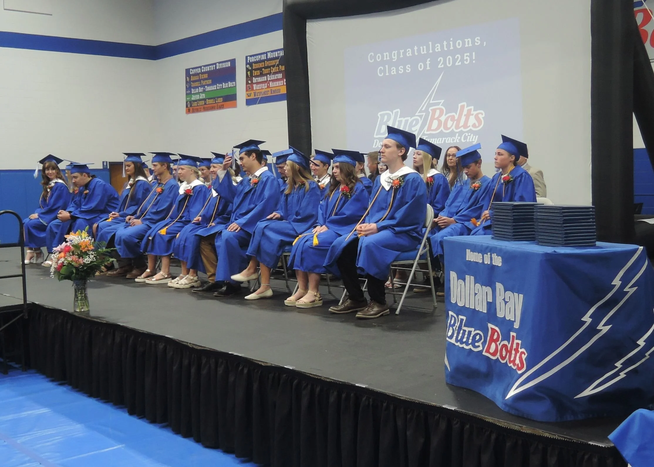 Picture of senior graduation.  A dozen seniors sitting in the front row and the others seated in the second row.  A table with diplomas stacked on it in the foreground.