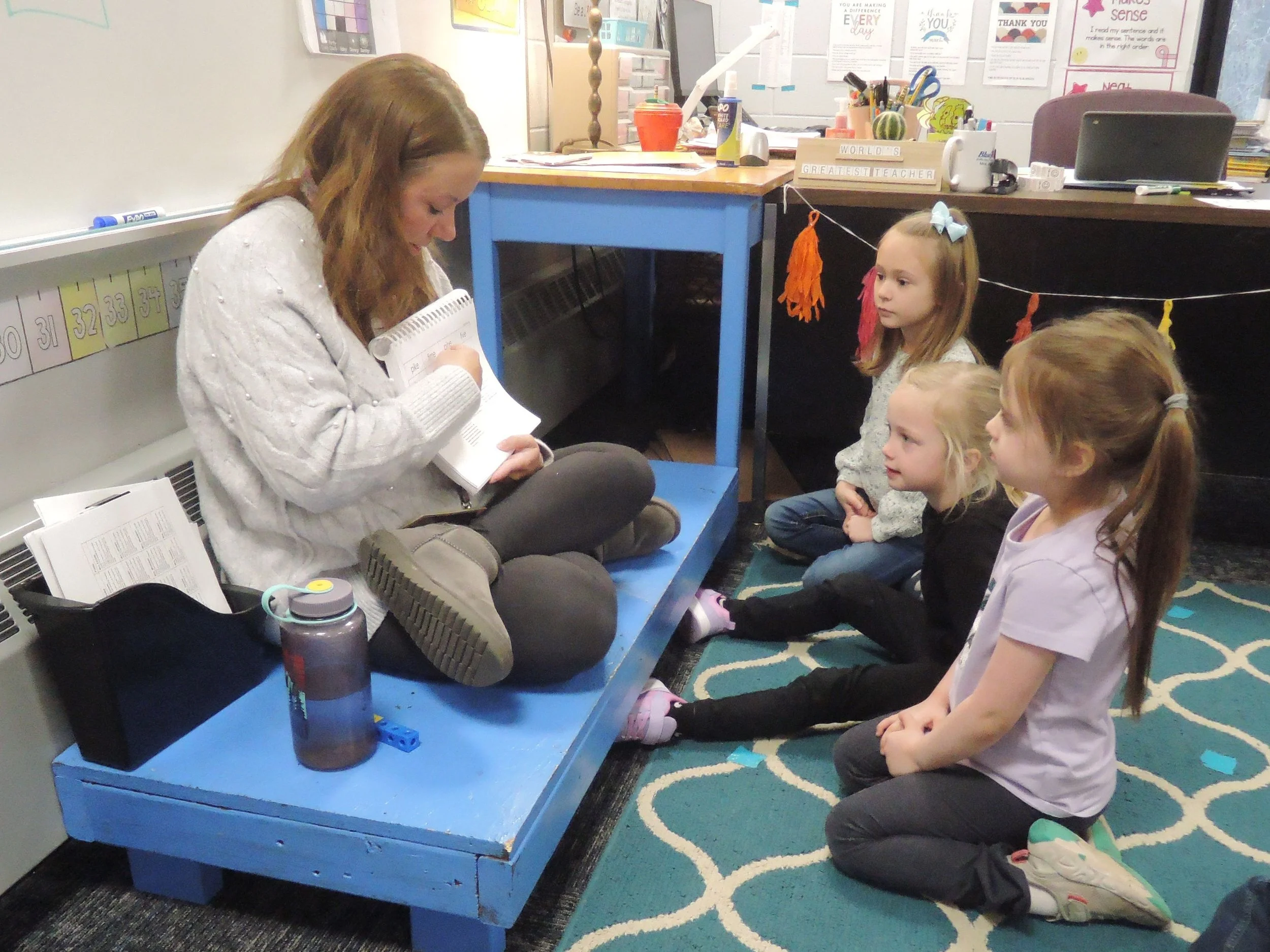 The kindergarten teacher sitting cross legged on a very short blue wooden platform, going over some language arts words with three student sitting on the carpeted floor in front of her.
