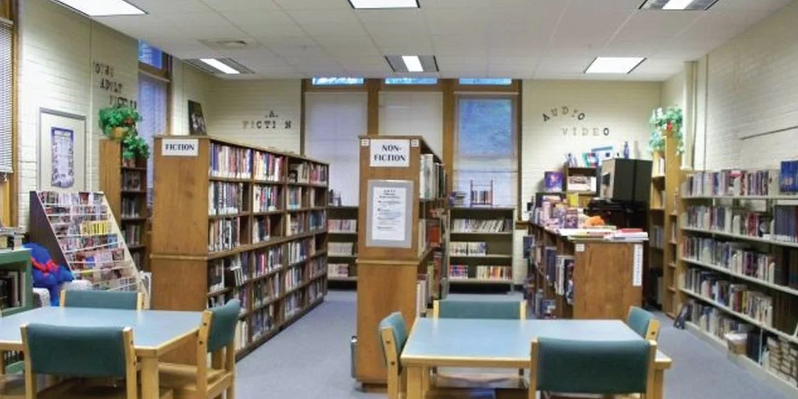 Picture of the high school wing of the library; book shelves along each wall and several bookshelves in the middle of the room.  There are attractive square tables in the foreground, each surrounded by chairs where people can sit and read.