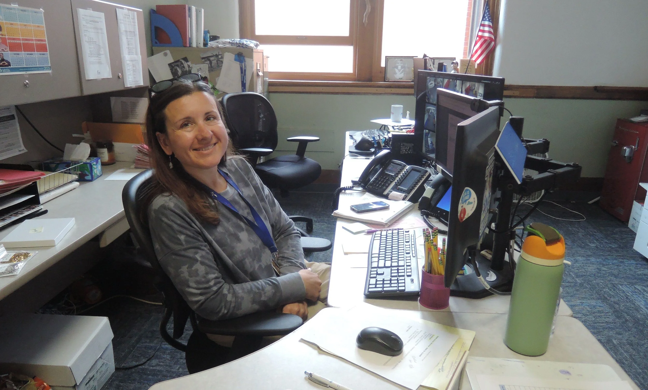 Side view picture of High School secretary at her desk, in front of a computer, smiling at the camera.
