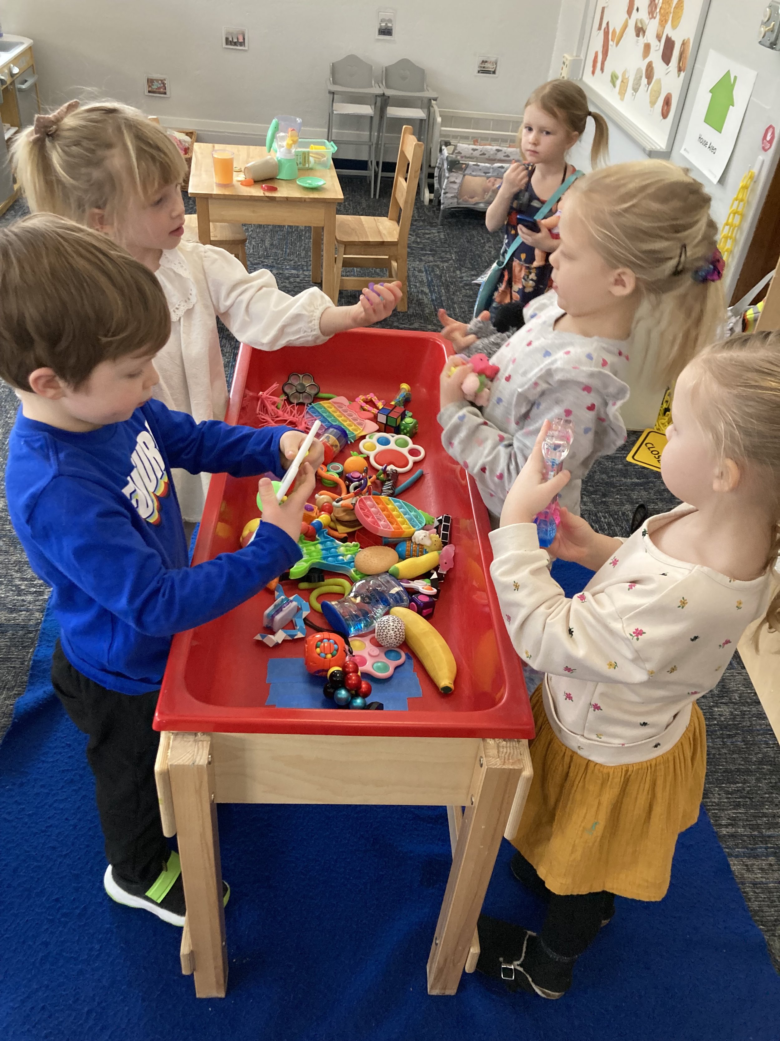 Four preschool students around a rectangular embedded table.  In the  embedded table are dozens of toy objects.s 