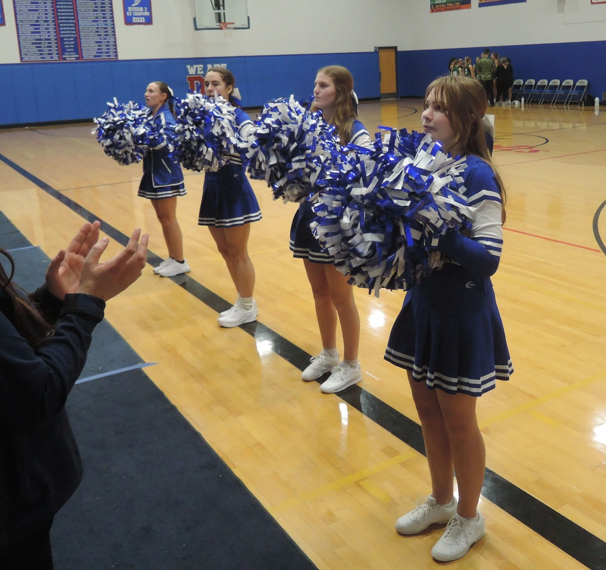 Four varsity cheerleaders, lined up on the side lines with pom poms, leading the fans in a cheer.