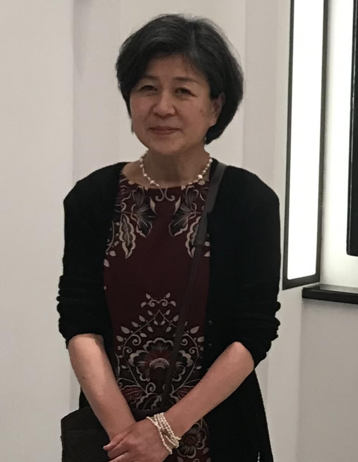 A middle-aged woman with short black hair, dressed in a patterned burgundy dress with black floral embroidery, wearing a pearl necklace and bracelet, standing indoors with a neutral expression.