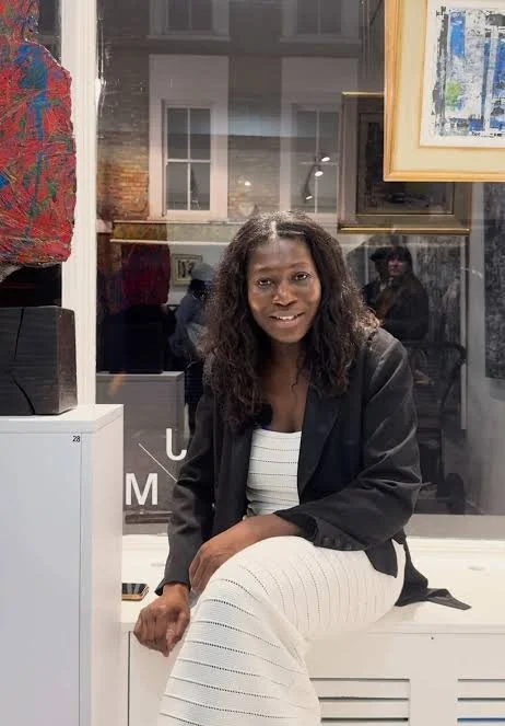 Woman with dark curly hair sitting on a white bench inside an art gallery, wearing a black blazer and white striped dress, with artwork displayed on the walls behind her.