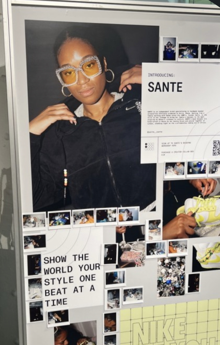 A woman wearing big rhinestone glasses and hoop earrings posing in front of a poster about Sante, with smaller photos and a quote about showing personal style, and images of her crafting and shoes on a display board.