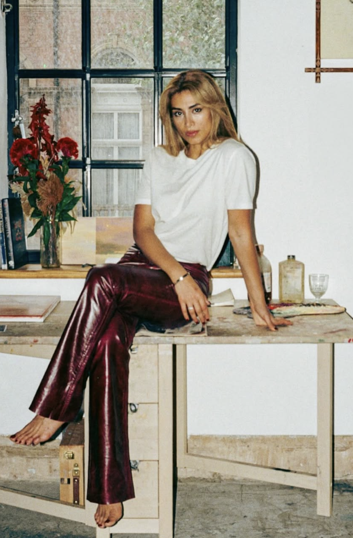 A woman with blonde hair wearing a white shirt and shiny red pants sitting on a rustic wooden table in a room with a large window. There are books, a vase with red flowers, bottles, and a glass on the table, and a map on the wall.