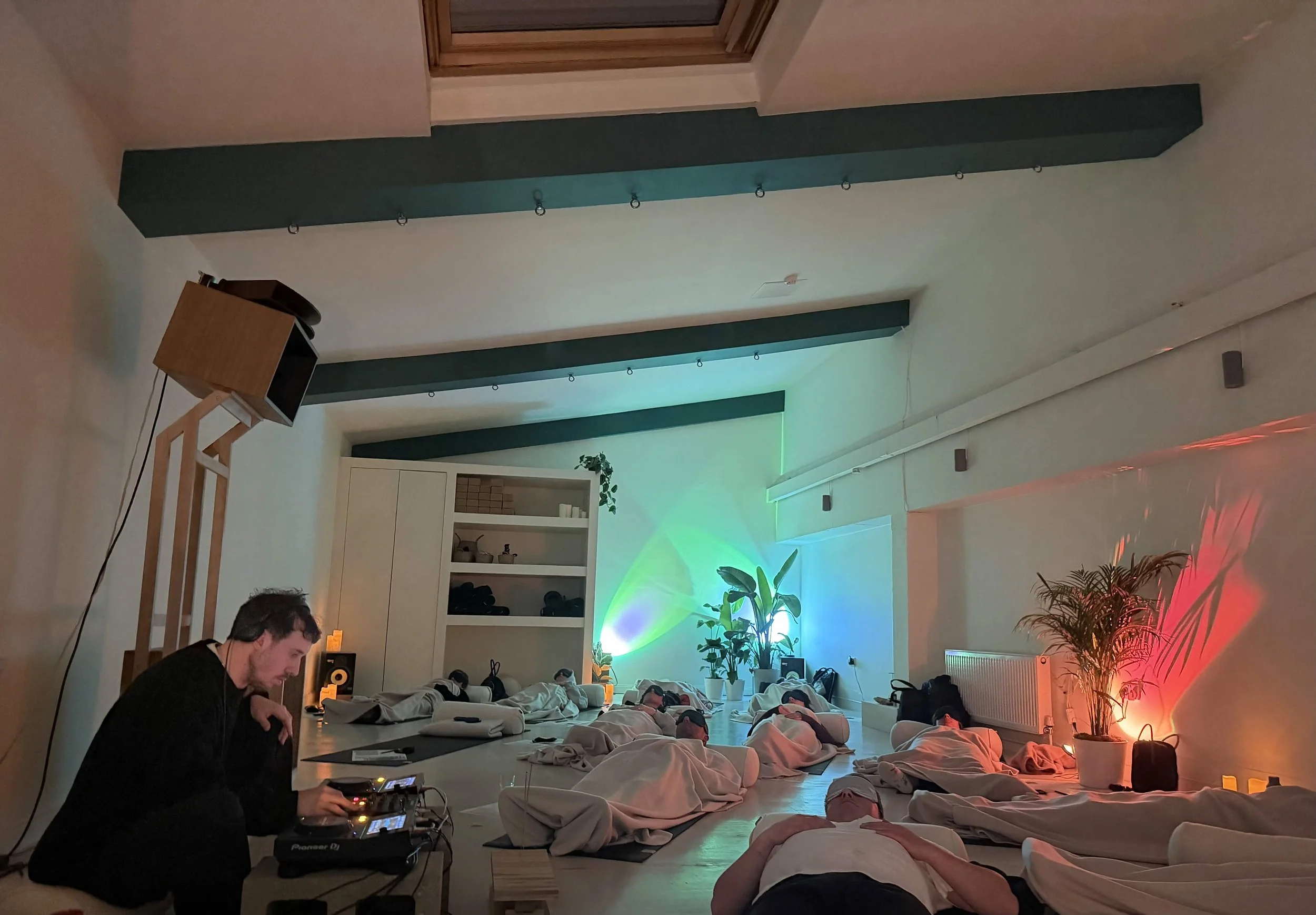 People lying on mattresses on the floor with their eyes closed, in a cozy room with colorful lighting and plants, while a DJ plays music.