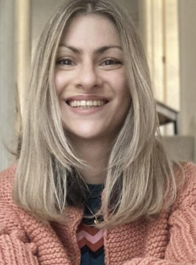 A young woman with long blonde hair smiling, wearing a pink knit sweater in a cozy indoor setting.