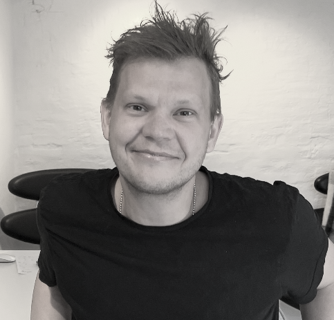 Ung mand med kort, lyst hår og smil, iført sort T-shirt og kæde, sidder i et rum med hvid væg og sportstole