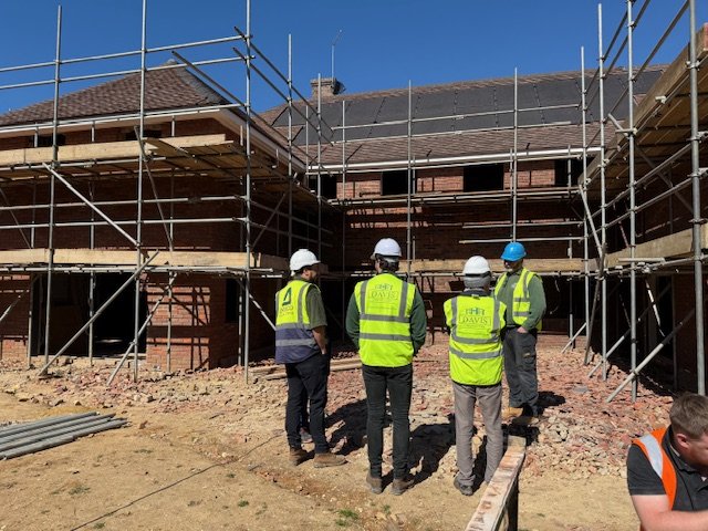 Group of construction workers wearing safety vests and helmets standing on dirt ground in front of a building under construction, surrounded by scaffolding.