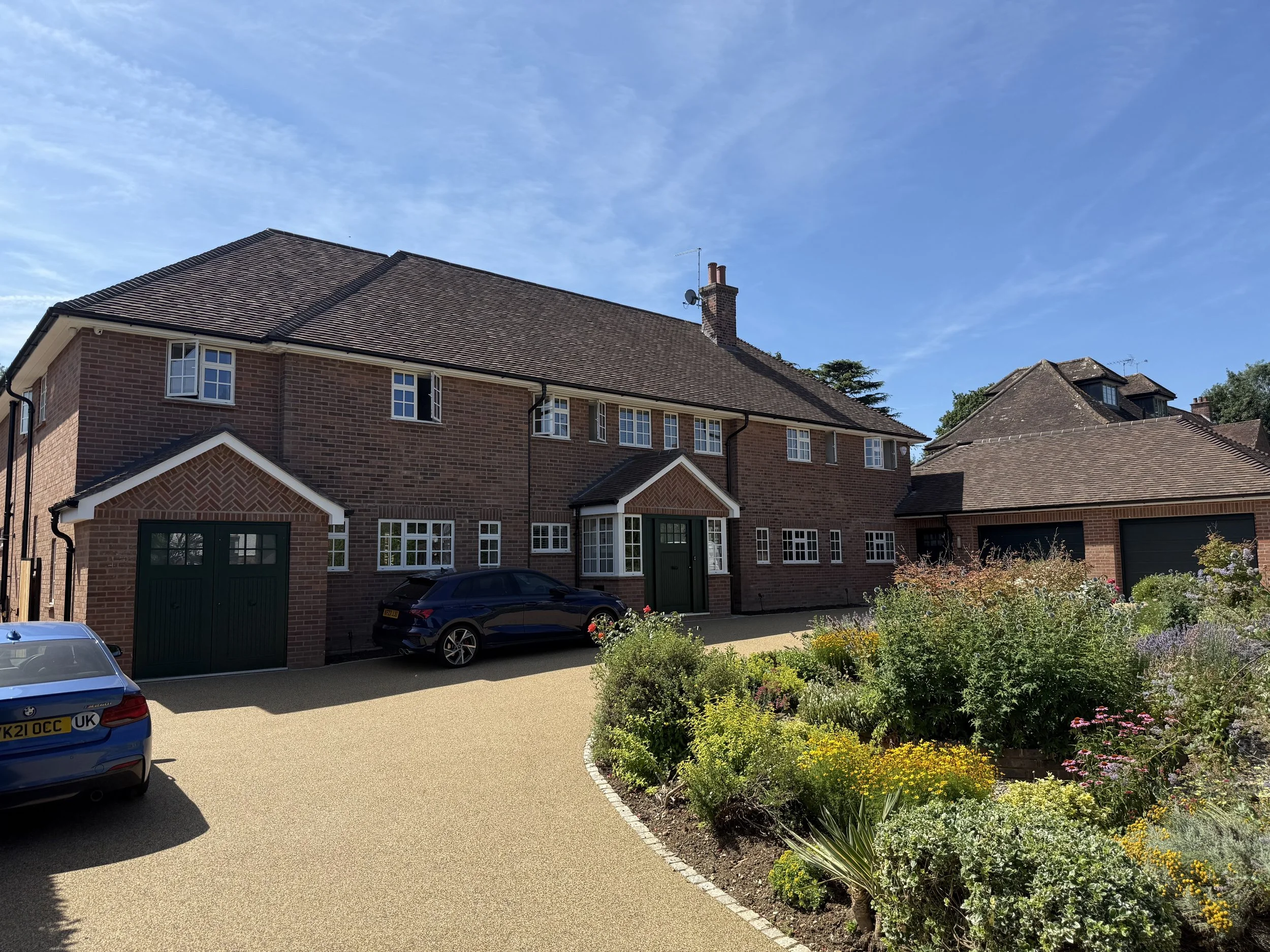 A row of brick houses with brown, tiled roofs and a front yard garden with colorful flowers, a large driveway, and parked cars under a blue sky with some clouds.