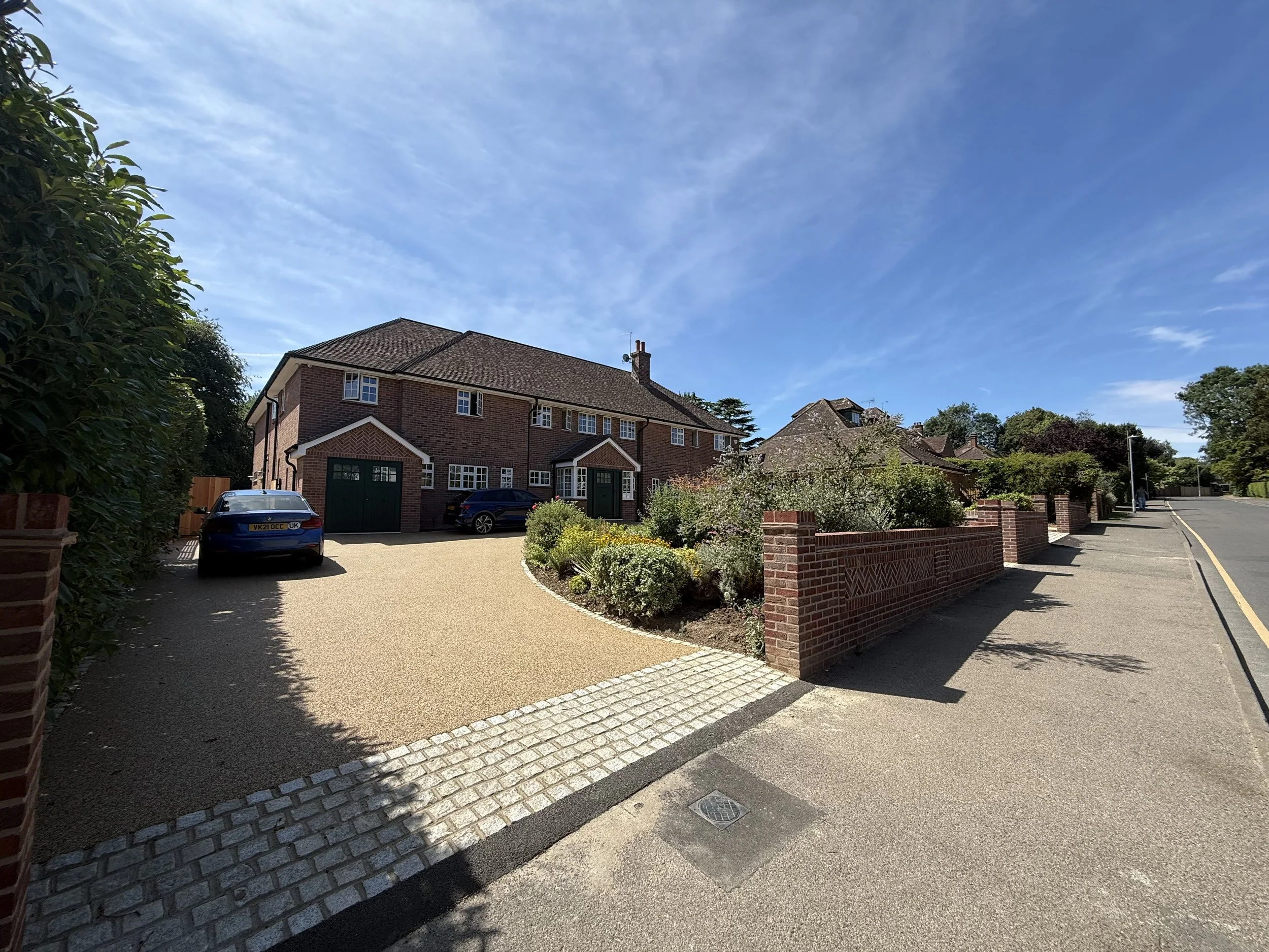 A residential neighborhood with brick houses, a driveway with a car, a garden with flowers, and a blue sky with some clouds.