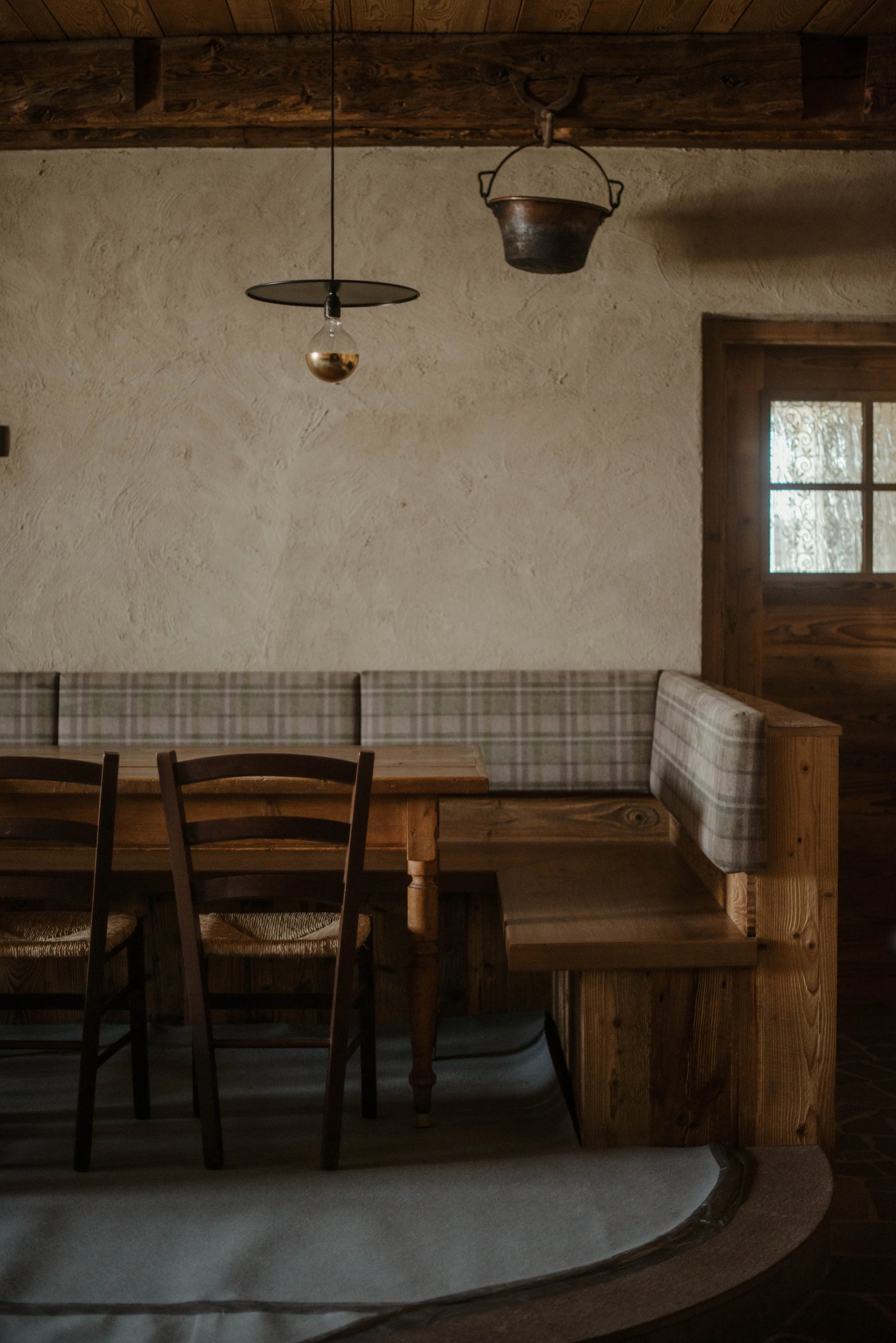 Angolo di una cucina rustica con parete in stucco chiaro, tavolo in legno a forma di L con sedie di legno e panche imbottite a scacchi, lampadario in metallo e finestra con tendina decorativa.