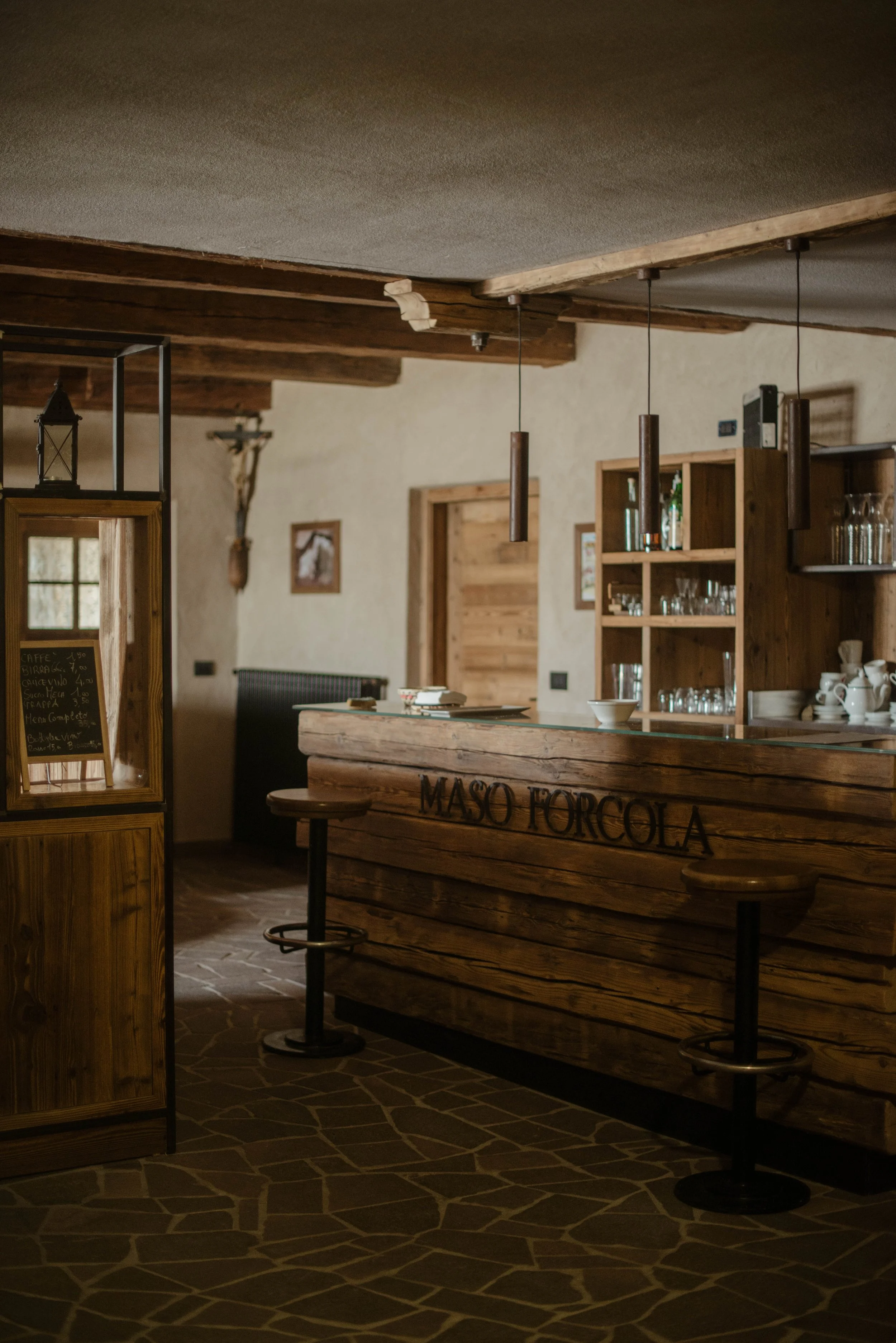 Interno di un bar rustico con banco in legno e sgabelli, scaffali con bicchieri e stoviglie, decorazioni in stile montano.