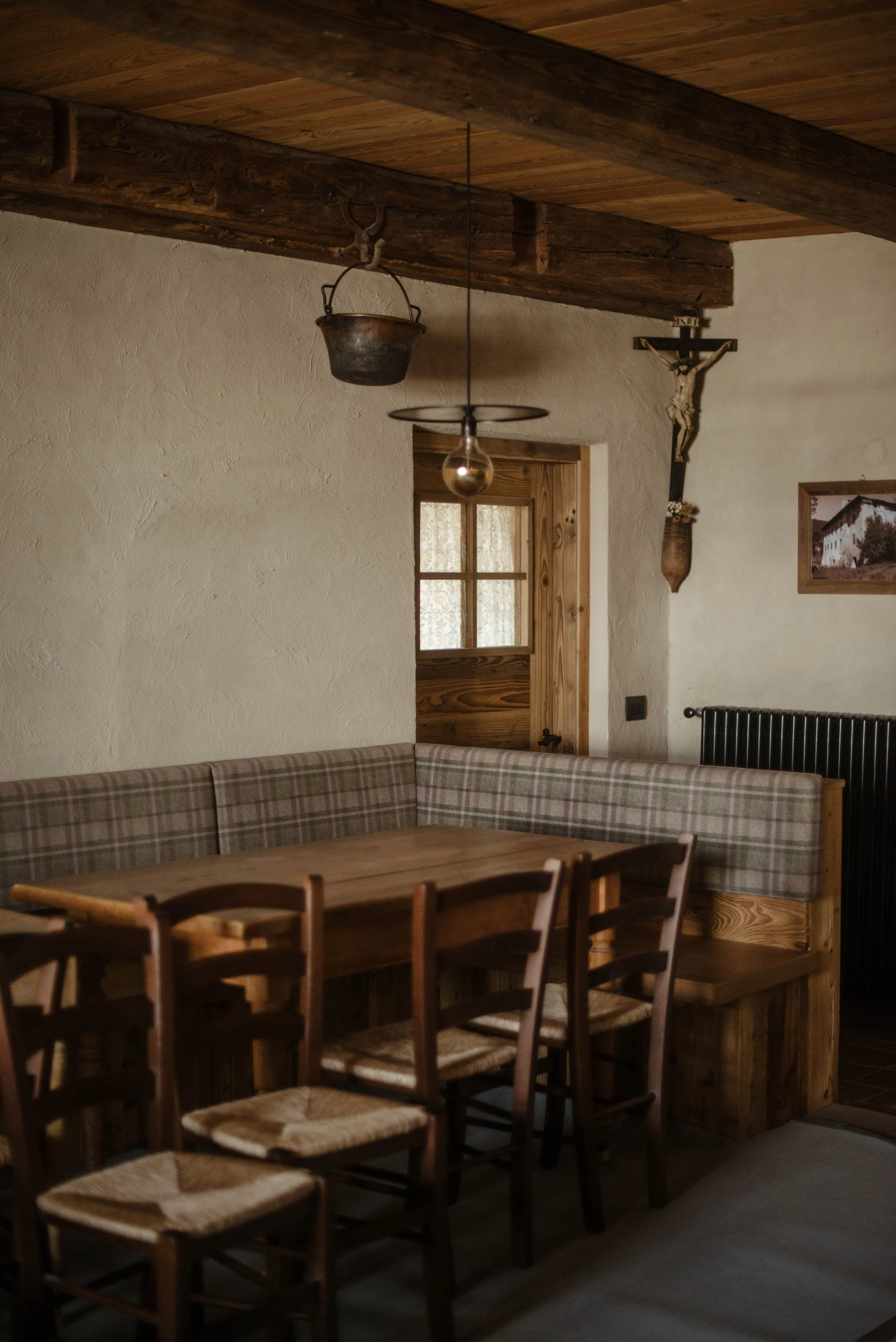 Interno di una stanza con tavolo di legno, sedie e panche in legno, pareti bianche, decorazioni rustiche e una finestra in legno.