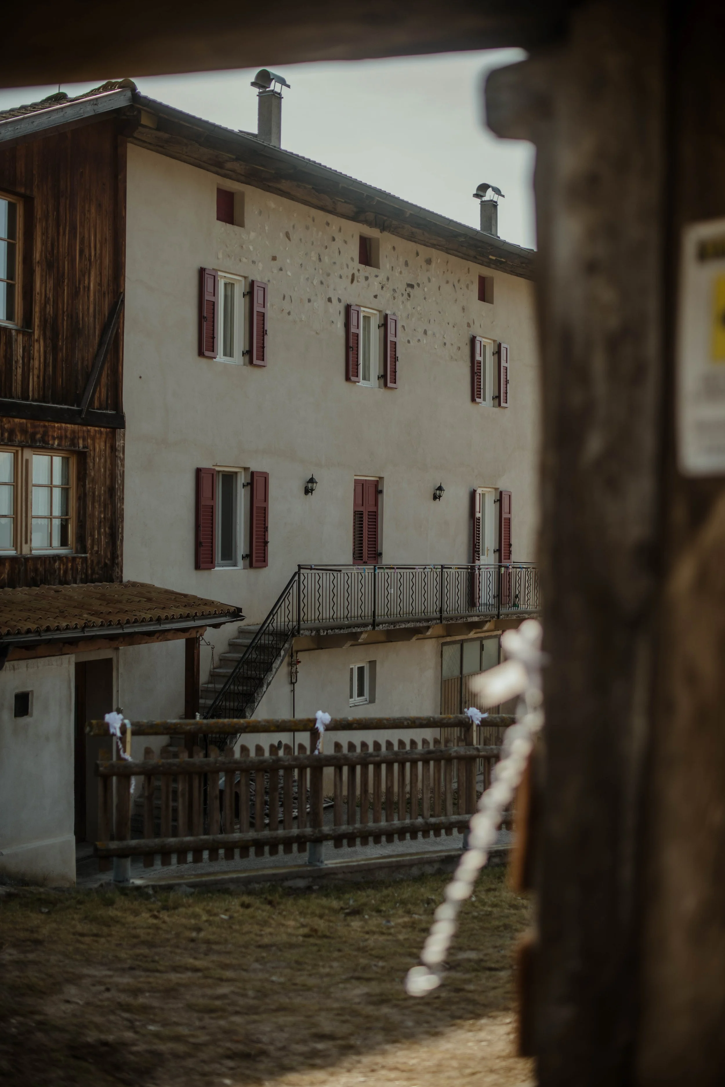 Vista di una casa in stile rustico con pareti chiare, finestre con persiane marroni e un balcone in ferro battuto, visibile da un'apertura nel legno.