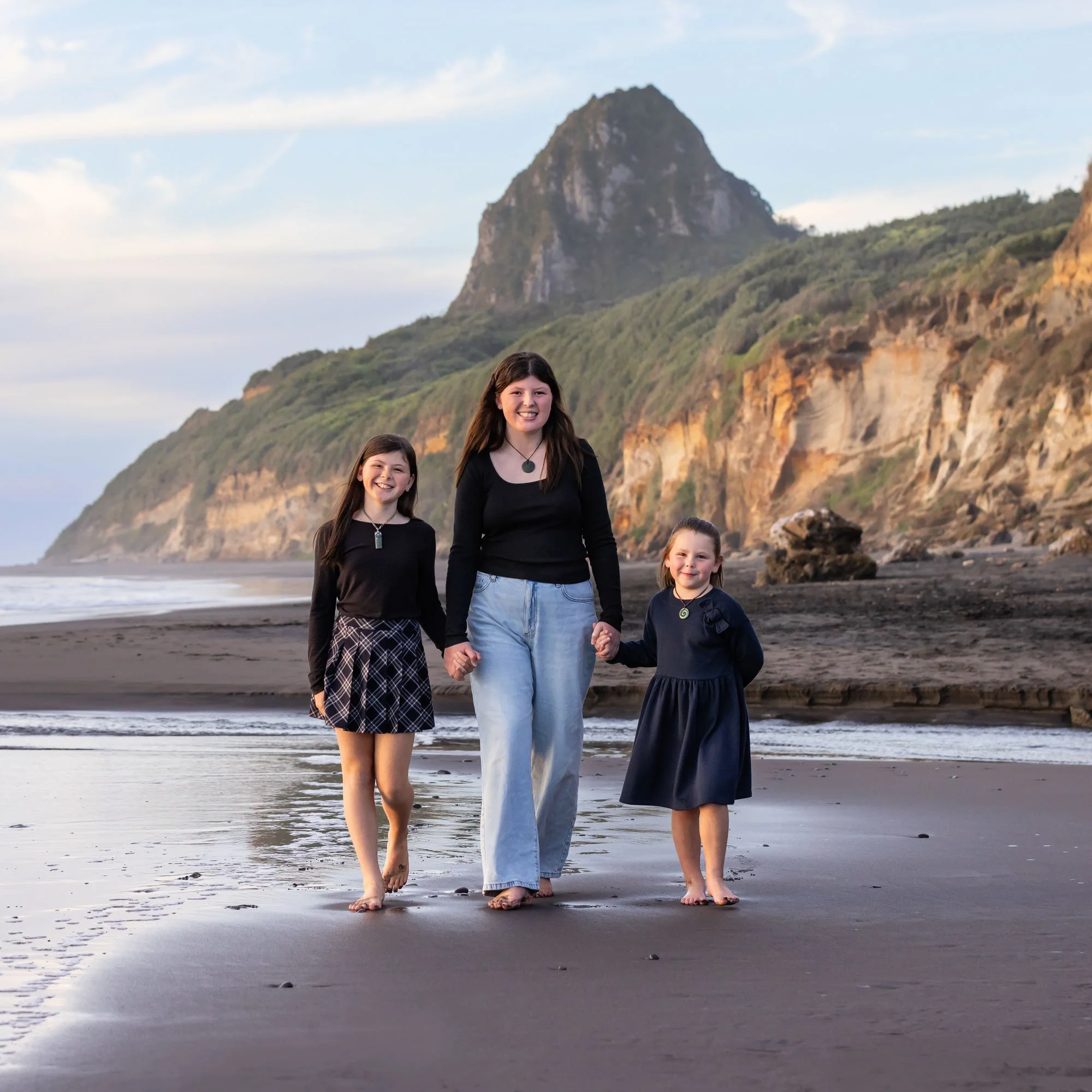 A woman and two young girls walking barefoot on a beach holding hands, with cliffs and a mountain in the background.