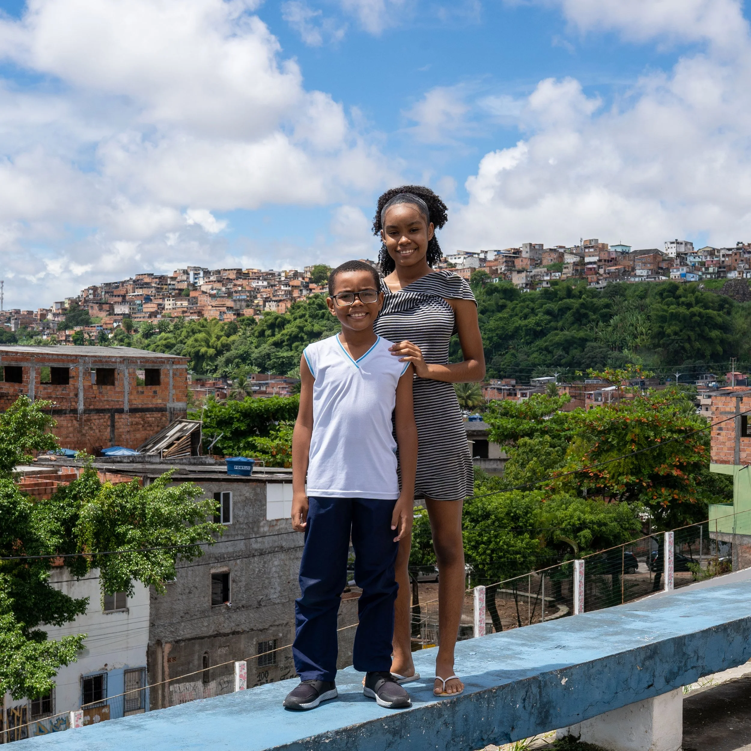 A boy and a girl standing on a concrete ledge outdoors with a hillside of densely packed houses in the background, under partly cloudy skies.