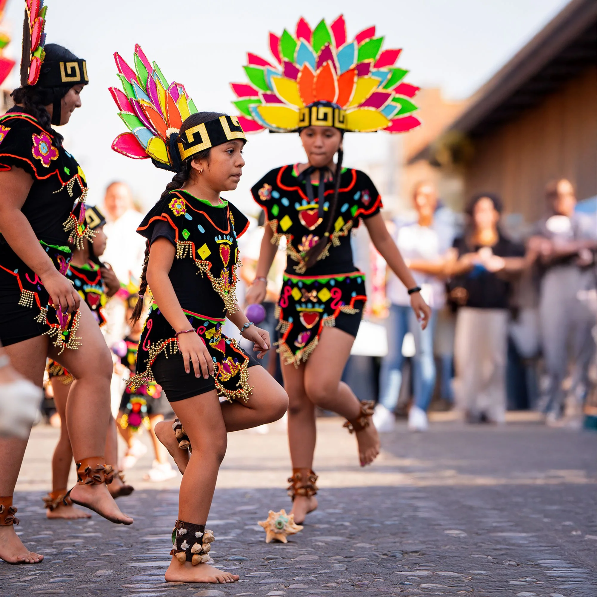 Children dressed in colorful traditional costumes participate in a cultural dance, wearing feathered headdresses and beaded accessories, on a cobblestone street.