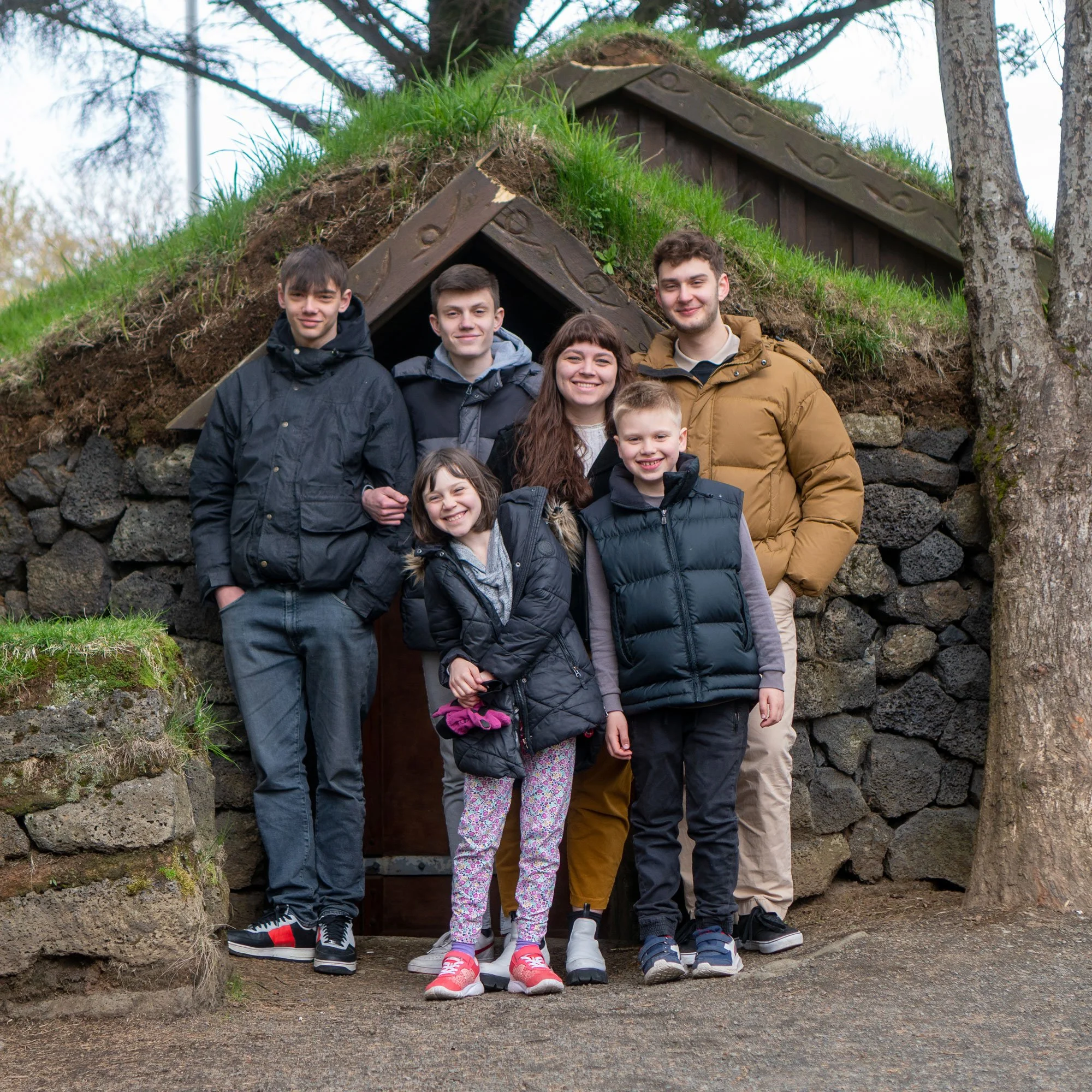 Group of six people, four children and two adults, standing in front of a small, grass-covered stone and wood structure outdoors.