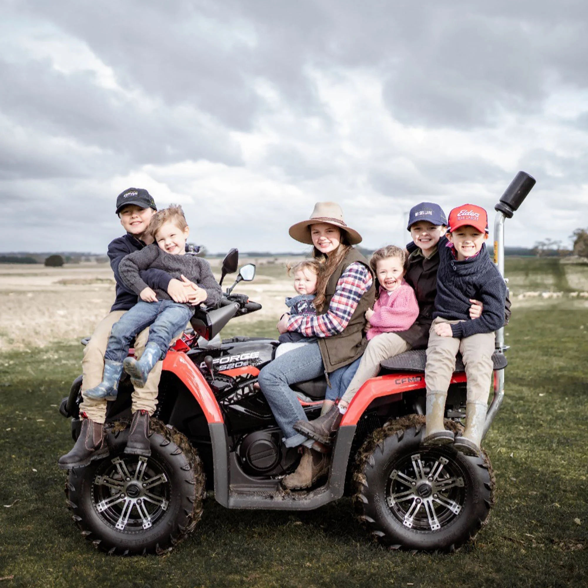 Group of children and a woman sitting on a red all-terrain vehicle (ATV) outdoors on a cloudy day, holding and hugging each other.