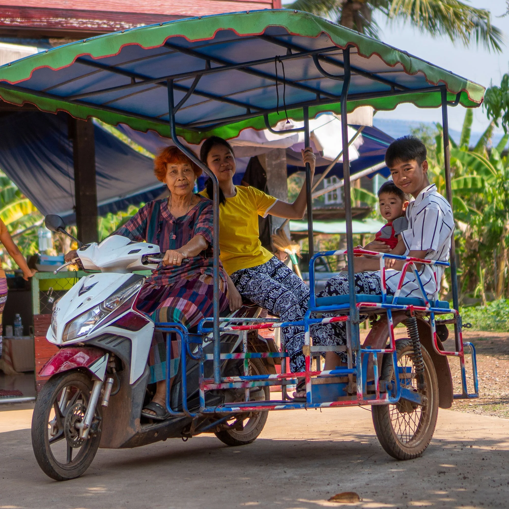 A family riding a motorized tricycle with a green canopy, with an older woman and two children on board, in a rural setting with trees and houses in the background.