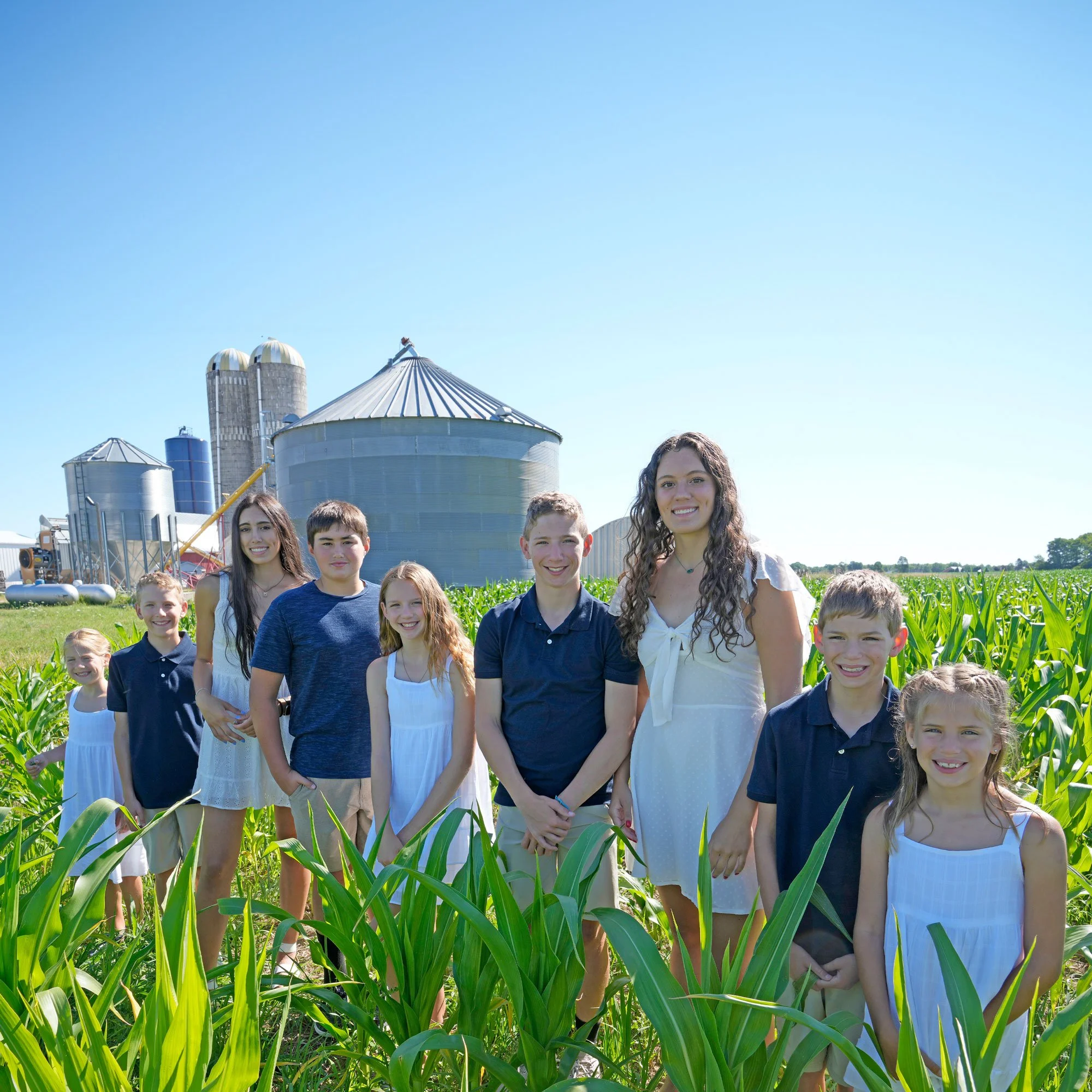 Group of children and teenagers standing in a cornfield with farm silos in the background on a bright, sunny day.