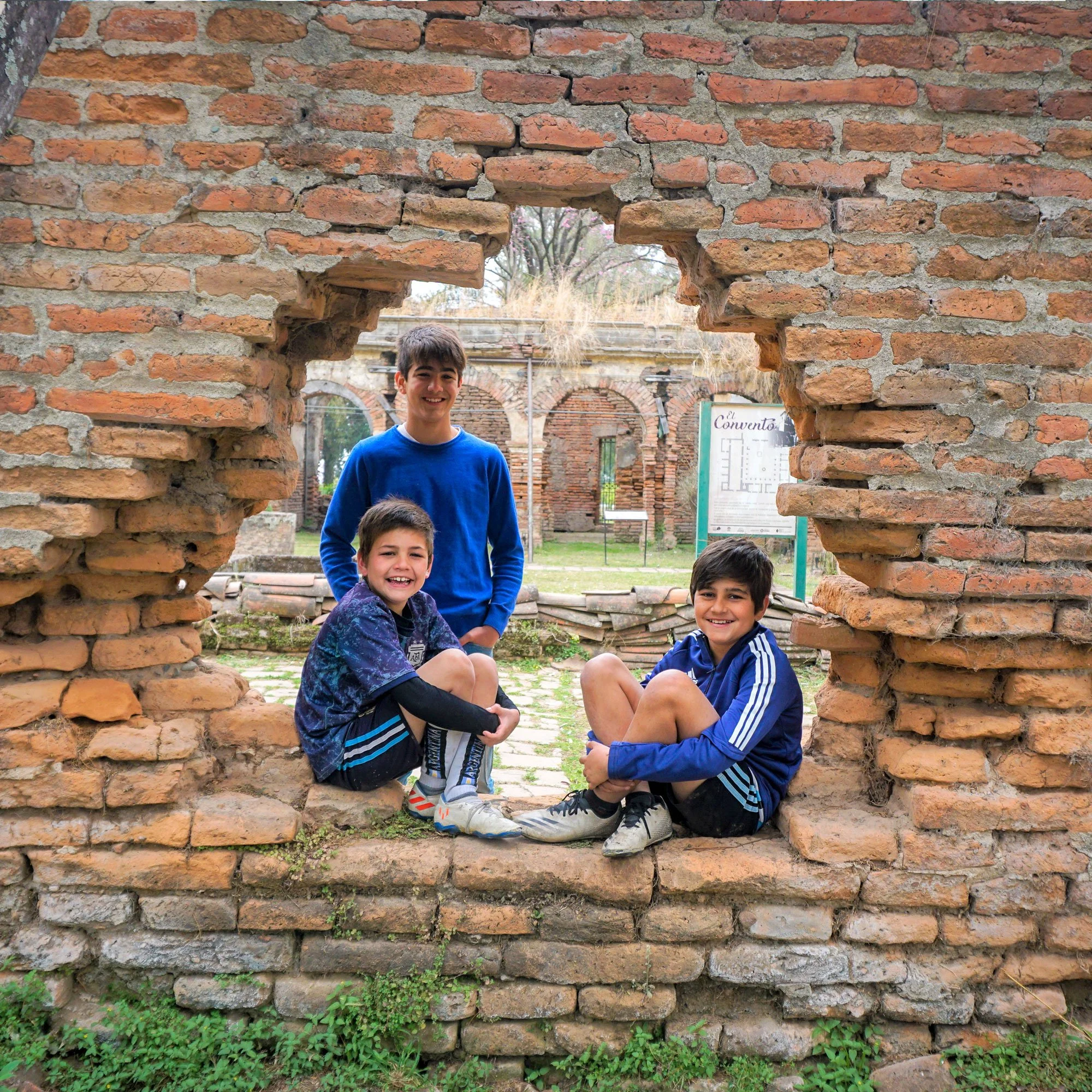 Three boys sitting and standing inside a large hole in a historic brick wall, smiling. Background shows more brick structures and a sign that reads 'Convento'.