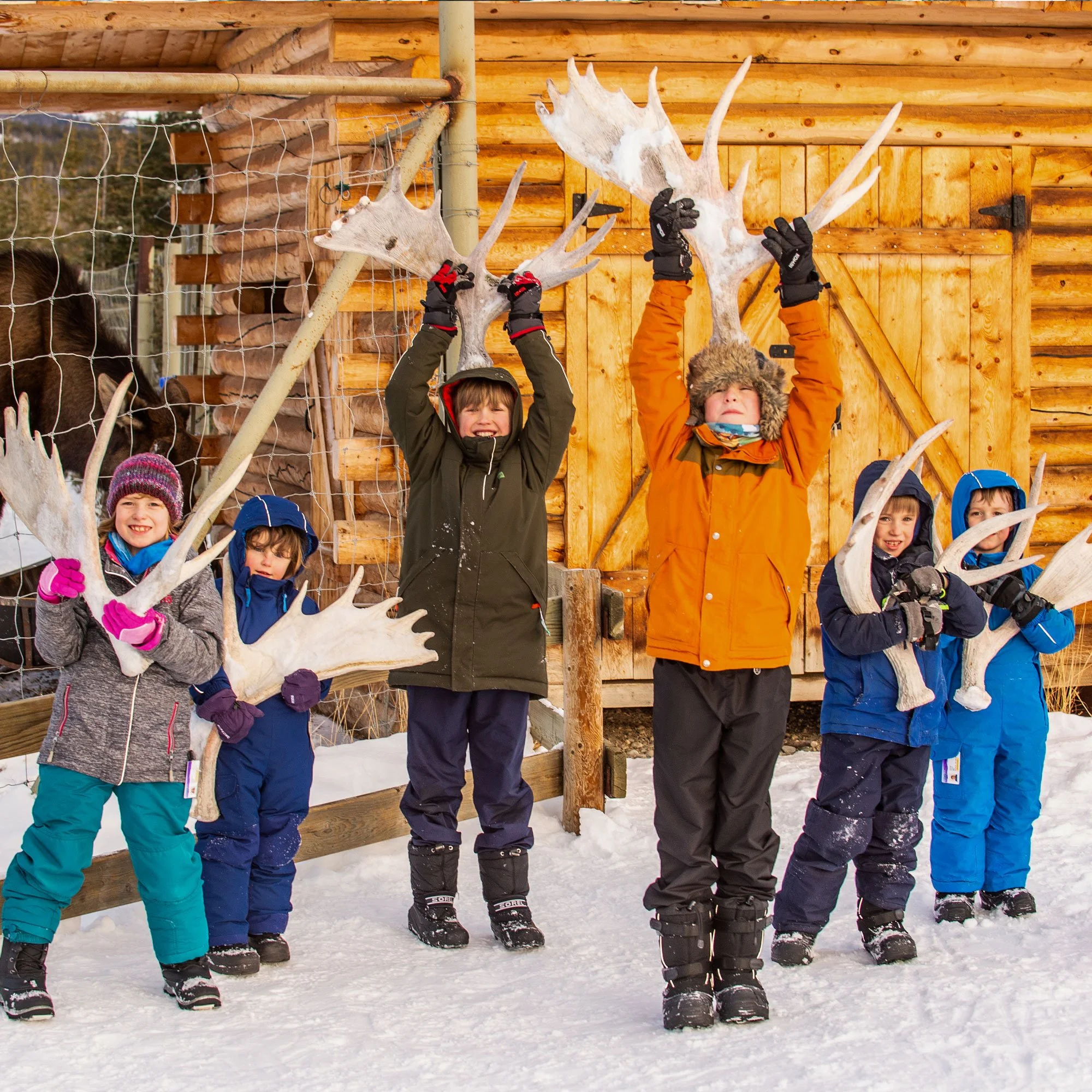 Six children in winter clothing holding large antlers in front of a wooden building and snow-covered ground.
