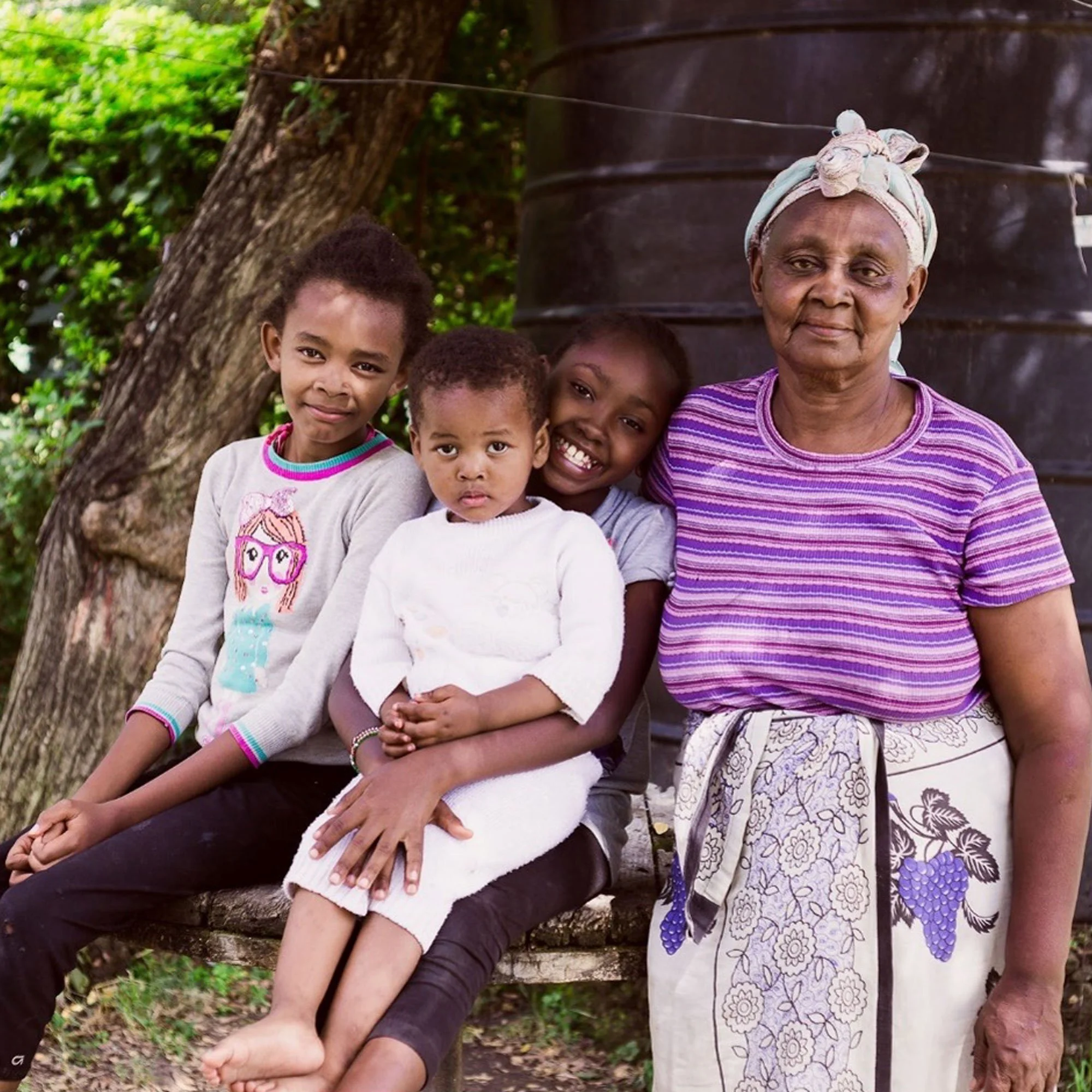 Four children and an elderly woman sitting on a wooden bench outdoors, with a large tree and a black water tank in the background.