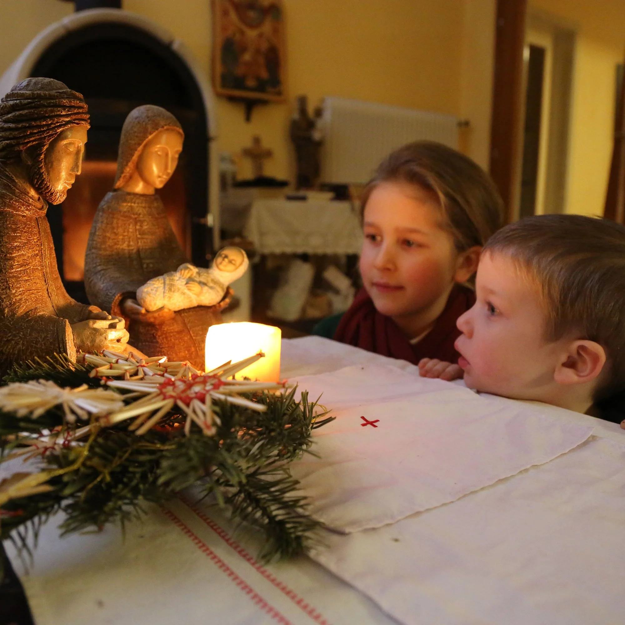 Two children observing nativity scene with candles and greenery on a table in a warmly lit room.
