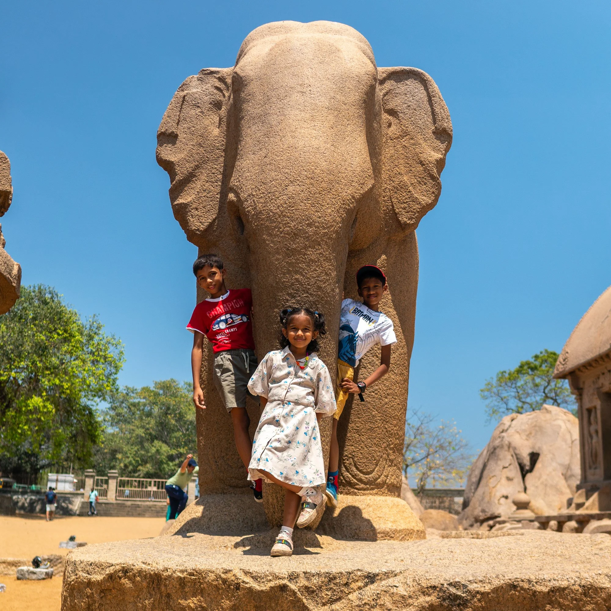 Three children standing in front of a large stone elephant sculpture outdoors on a sunny day.