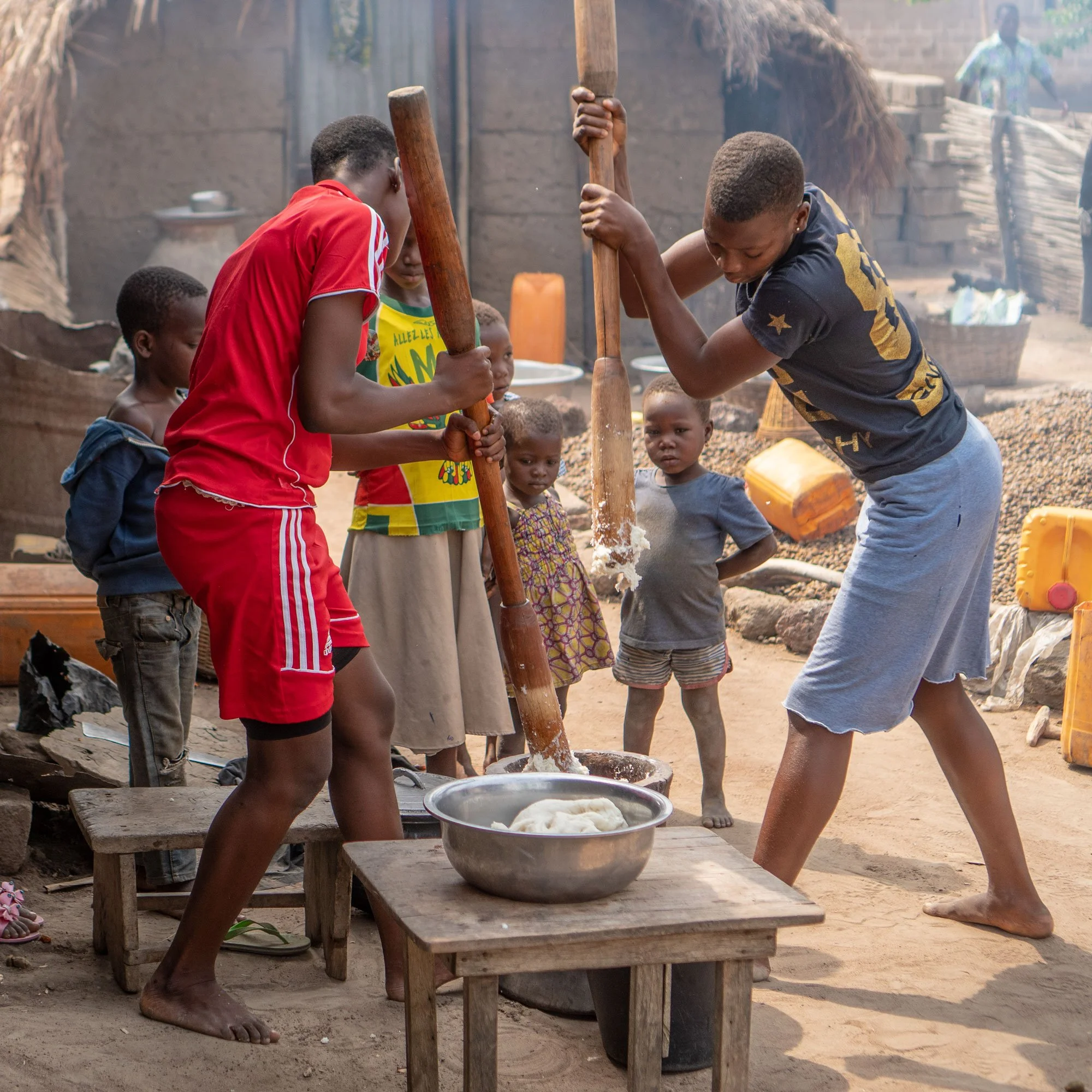 Two women are pounding maize with traditional sticks while children observe in a rural setting. There are various containers and a metal basin with dough on a wooden table nearby.