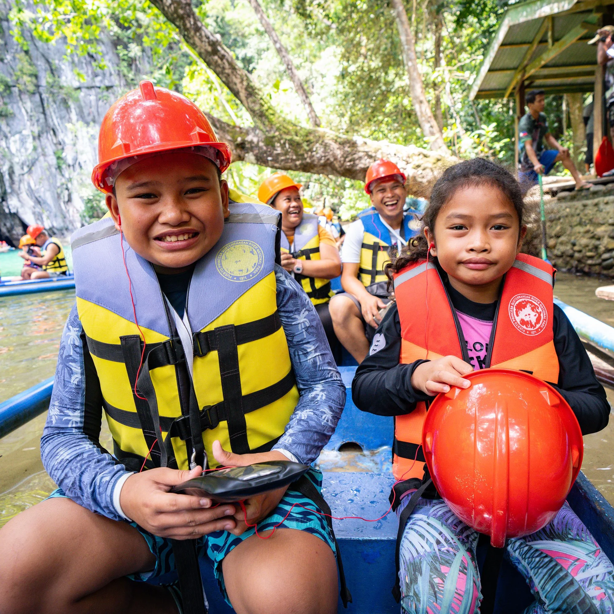 Children wearing life jackets and helmets on a boat during a river tour in a lush forested area.