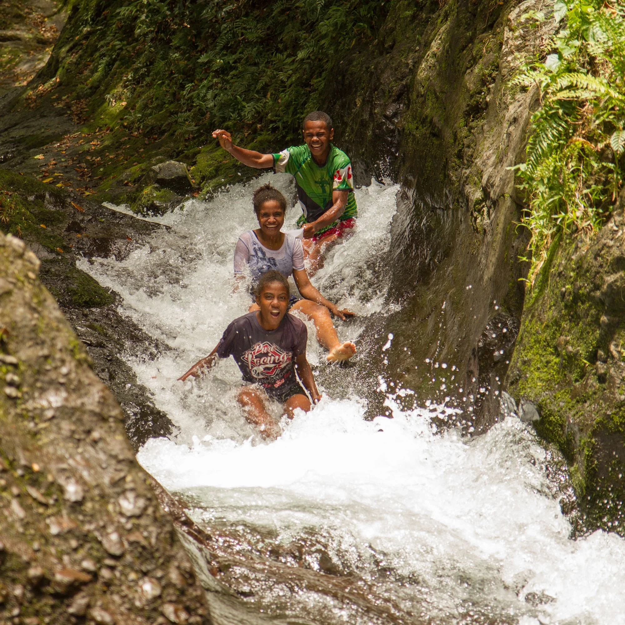 Four children playing and splashing in a narrow mountain stream, surrounded by lush green foliage and moss-covered rocks.