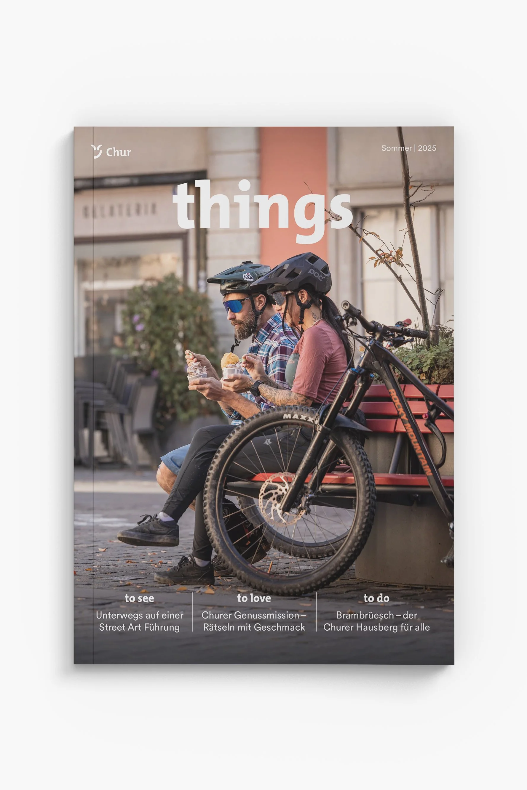 Ein Mann und eine Frau sitzen auf einer Parkbank, tragen Helme und essen Eis, mit einem mountainbike im Vordergrund, im Hintergrund eine Stadtstraße.