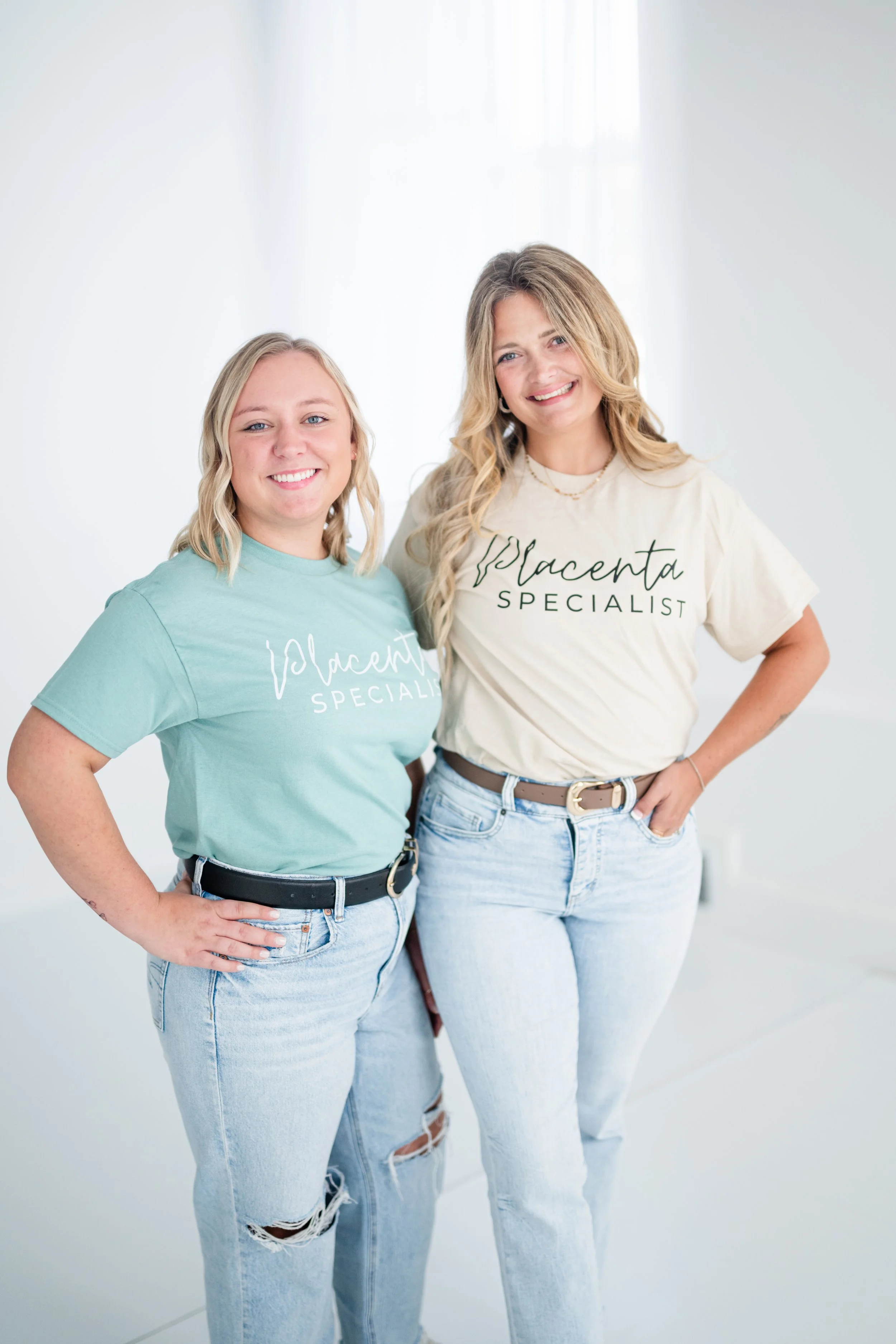 Two women wearing matching 'Placenta Specialist' t-shirts, standing in a bright, minimalistic room, smiling at the camera.