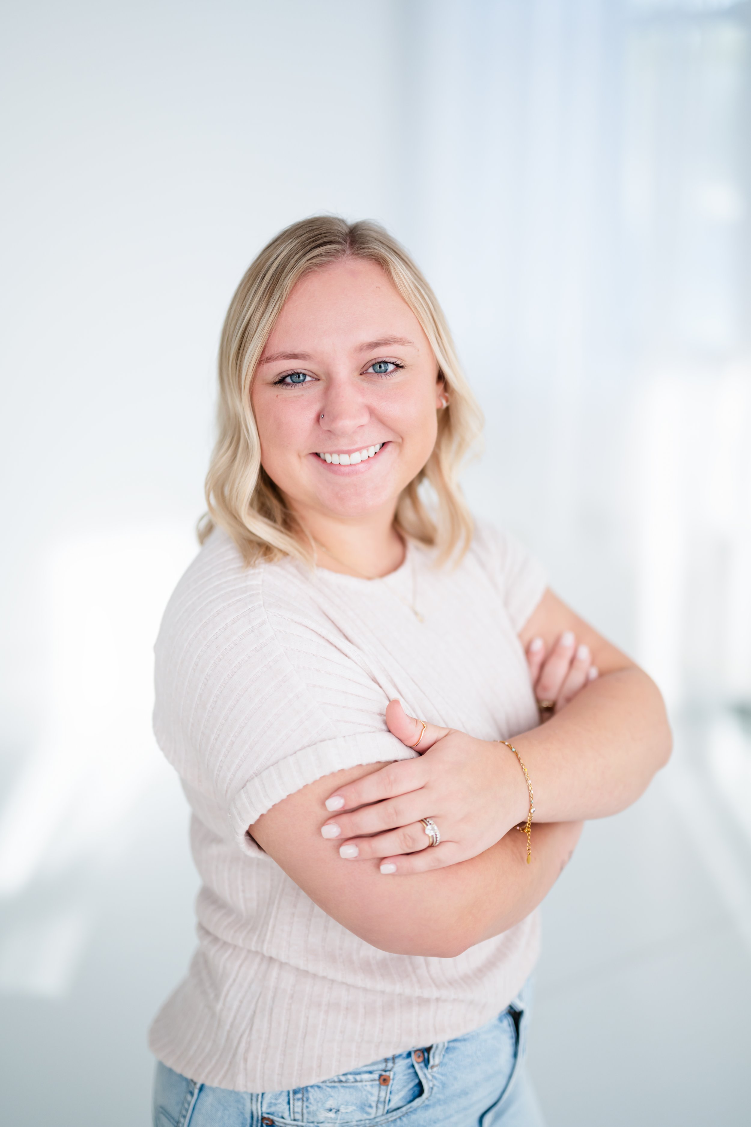 A young woman with blonde hair and blue eyes smiling, arms crossed, wearing a light-colored short sleeve shirt and jeans, in a bright, white room.