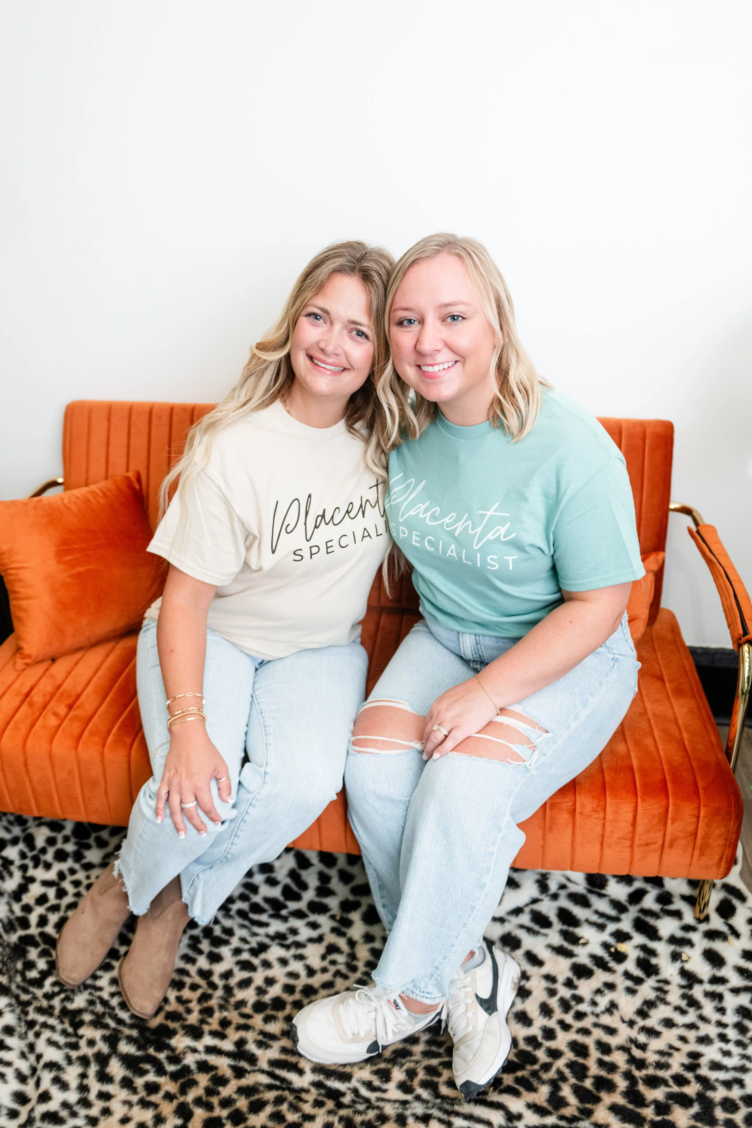 Two women sitting closely together on an orange velvet couch, smiling and posing for the camera. They are wearing casual t-shirts and jeans, with one woman wearing ripped jeans and sneakers, and the other wearing ankle boots. The woman on the left has blonde hair and is wearing a white t-shirt, and the woman on the right has blonde hair and is wearing a light teal t-shirt. The background is plain white with a leopard print rug on the floor.