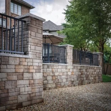 Exterior view of a stone retaining wall with black metal fence and a patio with umbrellas behind it.