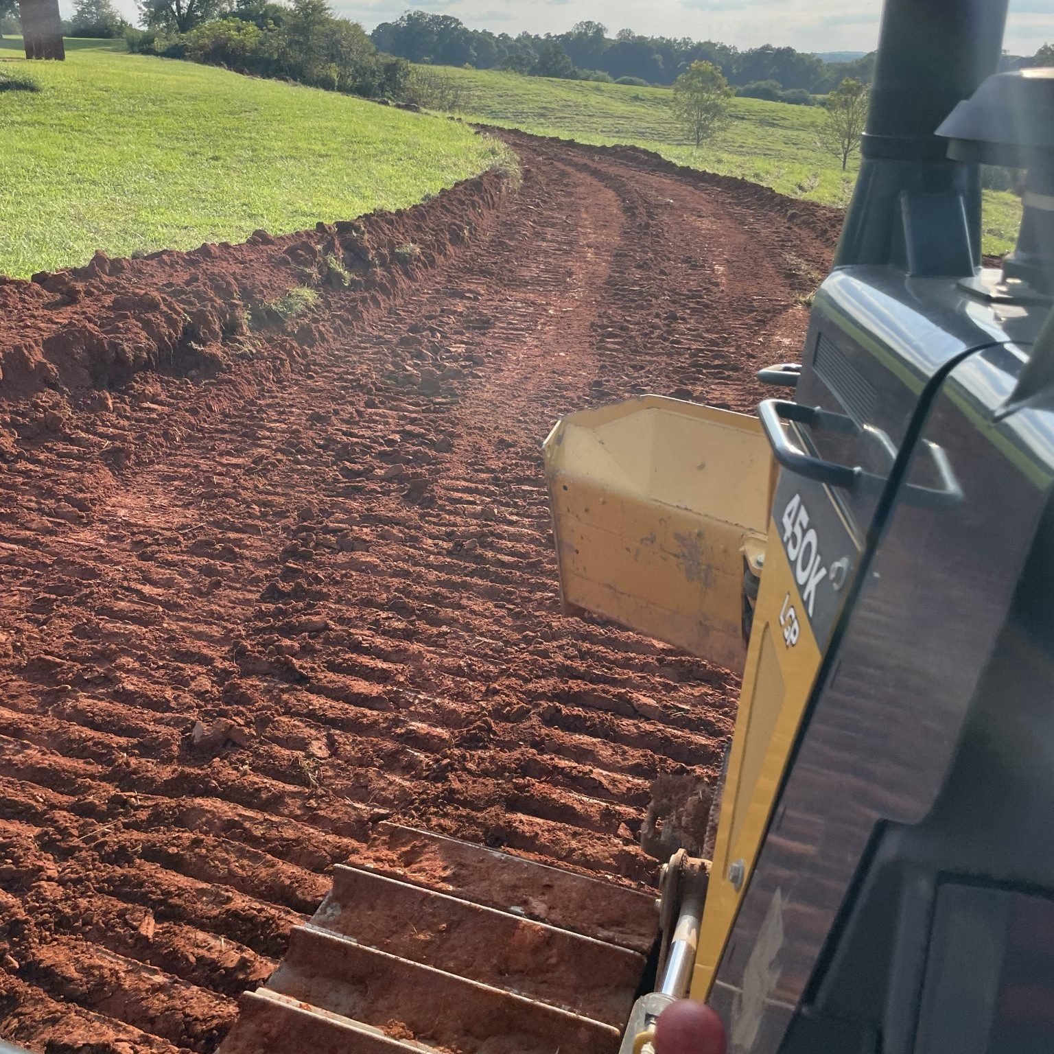 View from a tractor showing freshly plowed red dirt field on a farm, with green grassy areas and trees in the background.