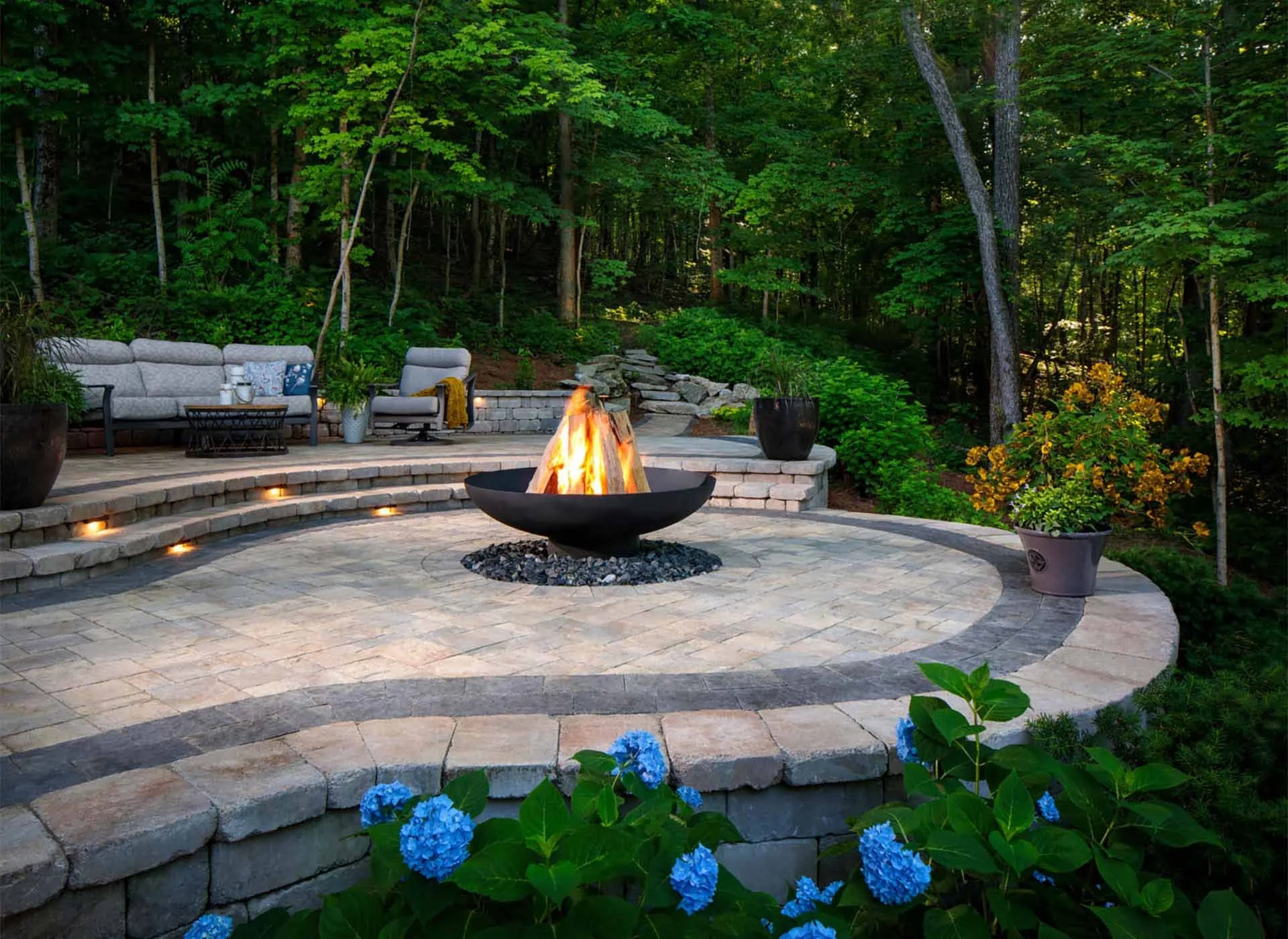 Outdoor backyard patio with a fire pit, outdoor furniture, potted plants, and surrounded by green trees and bushes.