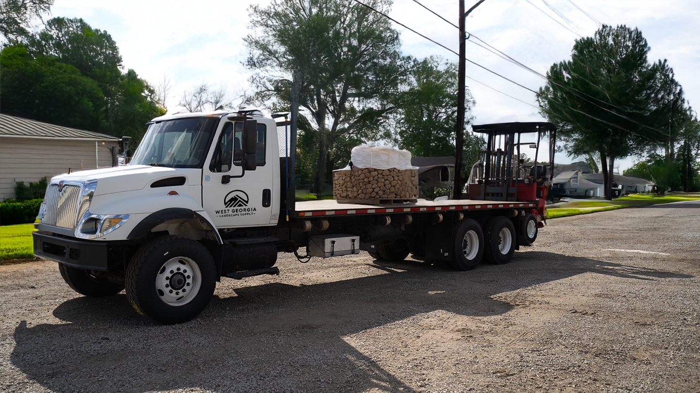 Flatbed truck with a forklift and bundled logs, marked with 'West Georgia Landscape Supply,' parked on a gravel lot with green trees and residential houses in the background.