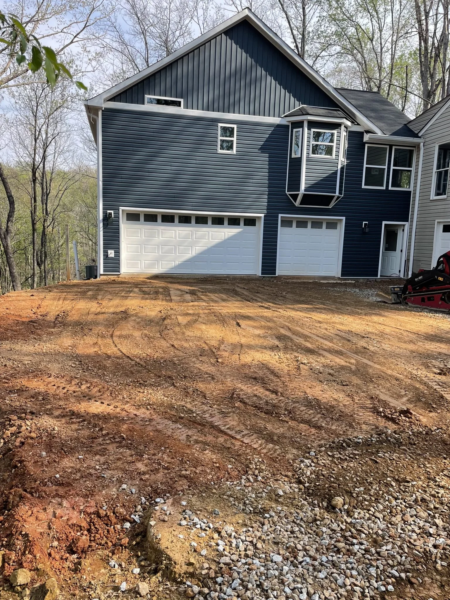 A newly constructed house with dark blue siding, white garage doors, and a small porch area. The front yard is still under construction with dirt and gravel, and a compact track loader is visible on the right side.